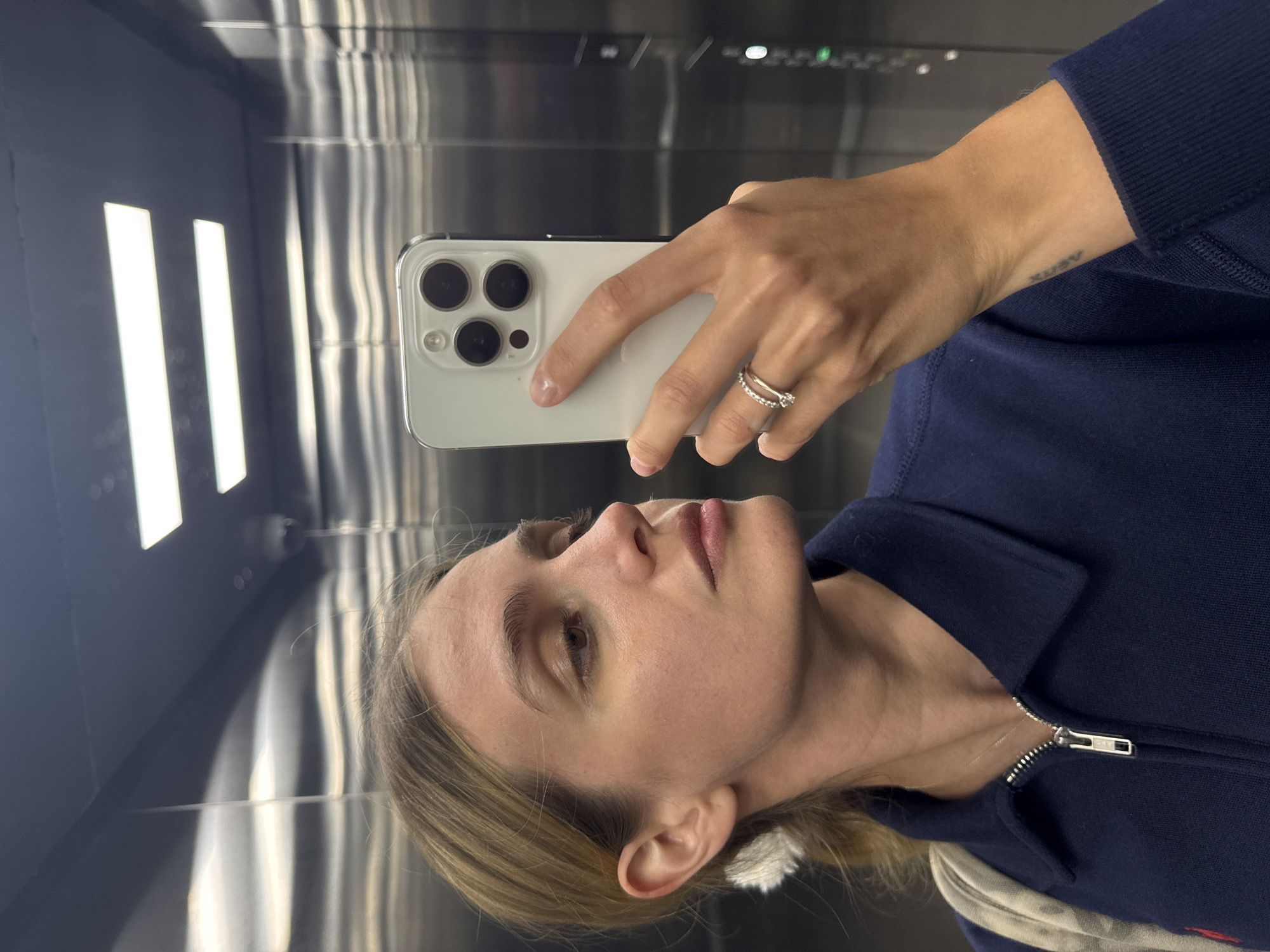A selfie taken in an elevator with stainless steel walls. A woman is holding a white smartphone in front of her face. She has blonde hair and is wearing a dark blue jacket with a zipper. She has a ring on her finger and a small tattoo on her wrist. The location is Unknown, Unknown.