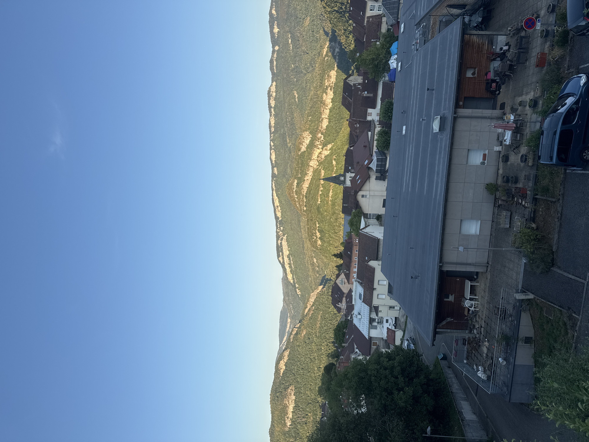 This is a high-angle, eye-level, medium shot of a village nestled in a valley with mountains in the background, located in Lavans-lès-Saint-Claude, France. The sky is a clear, light blue with a faint wispy cloud visible in the upper right quadrant. The foreground features a large, dark, flat-roofed building with some architectural details like skylights and vents. Adjacent to this building is a paved outdoor area with several chairs and tables, one with a partially closed umbrella, and various potted plants. A dark blue minivan is parked on the left side of the frame in front of a gray retaining wall and a road. Beyond this building, the village is visible with numerous houses with red-tiled roofs. A prominent church steeple with a clock face rises above the houses in the mid-ground. The background is dominated by lush green, forested mountains with distinct rocky escarpments, illuminated by the soft light of what appears to be early morning or late afternoon sun. The overall scene conveys a peaceful, semi-rural atmosphere.