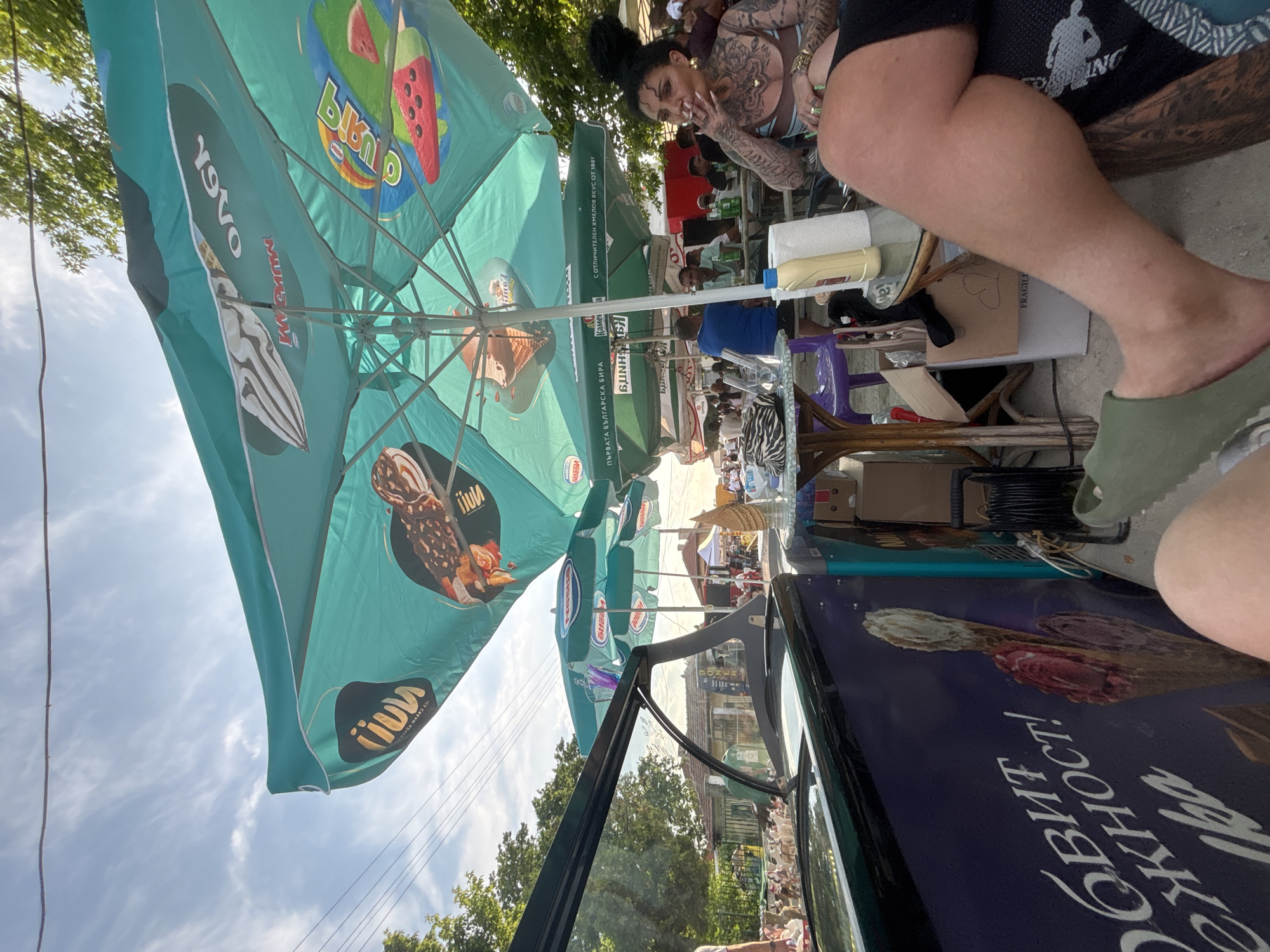 The image depicts an outdoor cafe scene in Kostievo, Bulgaria, under a bright, sunny sky with scattered cirrus clouds and visible power lines. The perspective is from a seated individual, with their bare legs, wearing an olive green slide sandal on the left foot, and extensively tattooed right leg (wearing dark, patterned shorts) visible in the foreground.
To the immediate left, a dark-colored ice cream display freezer is prominent, featuring Cyrillic text partially reading "ОБИТ НЕЖНОСТ!" (likely "Abundance of Tenderness!") alongside a large graphic of three ice cream cones. In the mid-ground, a series of teal-colored outdoor parasols dominate the upper frame, providing shade. These parasols bear various inverted ice cream brand logos, including "YAVA," "NESTLE," and "BINU," as well as a beer advertisement in Cyrillic, "ПЪРВАТА БЪЛГАРСКА БИРА КАМЕНИЦА" (The First Bulgarian Beer Kamenitza).
Underneath these parasols, multiple patrons are seated at various tables. To the right, a woman with dark hair styled in a bun, visible neck, arm, and chest tattoos, is seated, exhaling smoke from a cigarette. She wears a light-colored, possibly striped, top. On the table next to her are a yellow squeeze bottle and a roll of paper towels. Other individuals, primarily men, are visible seated at nearby tables in the background.
Beyond the immediate seating area, the scene extends to a street or open plaza. On the left, green foliage from trees is visible, with distant buildings, some appearing older with traditional architecture, and a sign partially visible on one building reading "ДОНЕ." The overall environment suggests a lively, casual outdoor gathering spot in a small town or village.