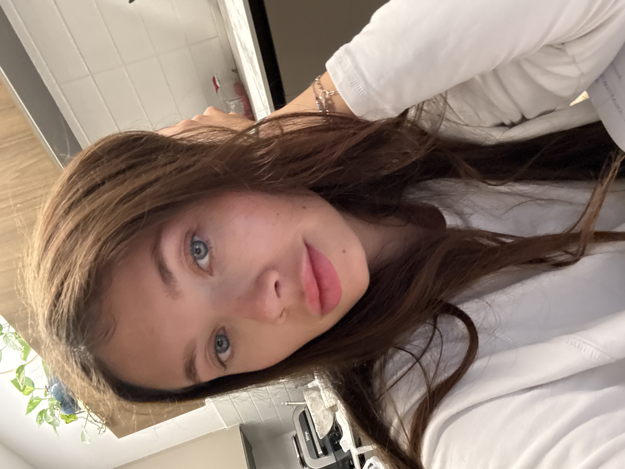 A close-up, slightly angled selfie photograph shows a young woman with long brown hair and blue eyes. She is wearing a white long-sleeved shirt and a silver bracelet is visible on her wrist. The background appears to be a kitchen or bathroom setting with white tiled walls and cabinets. A small plant is visible in the lower left corner. A Ninja blender is on the counter. The location is not apparent from the image and therefore cannot be said to be in Tel-Aviv, Israel.