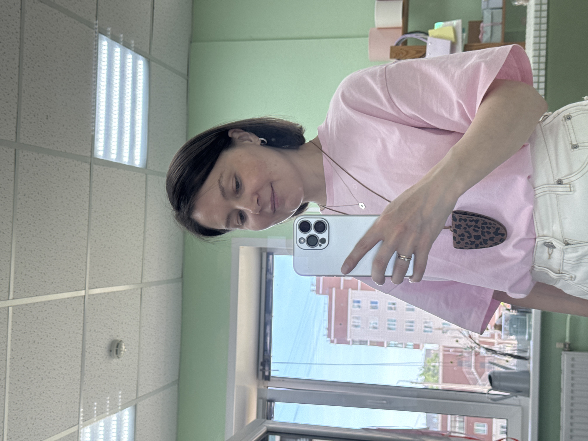 A woman takes a selfie in a room with a suspended grid ceiling containing fluorescent lights. She is wearing a pink t-shirt and white denim overalls. She is holding a white smartphone. Outside the window, a building can be seen. Based on the architecture and the surrounding environment visible through the window, the location is in Izhevsk, Russia.