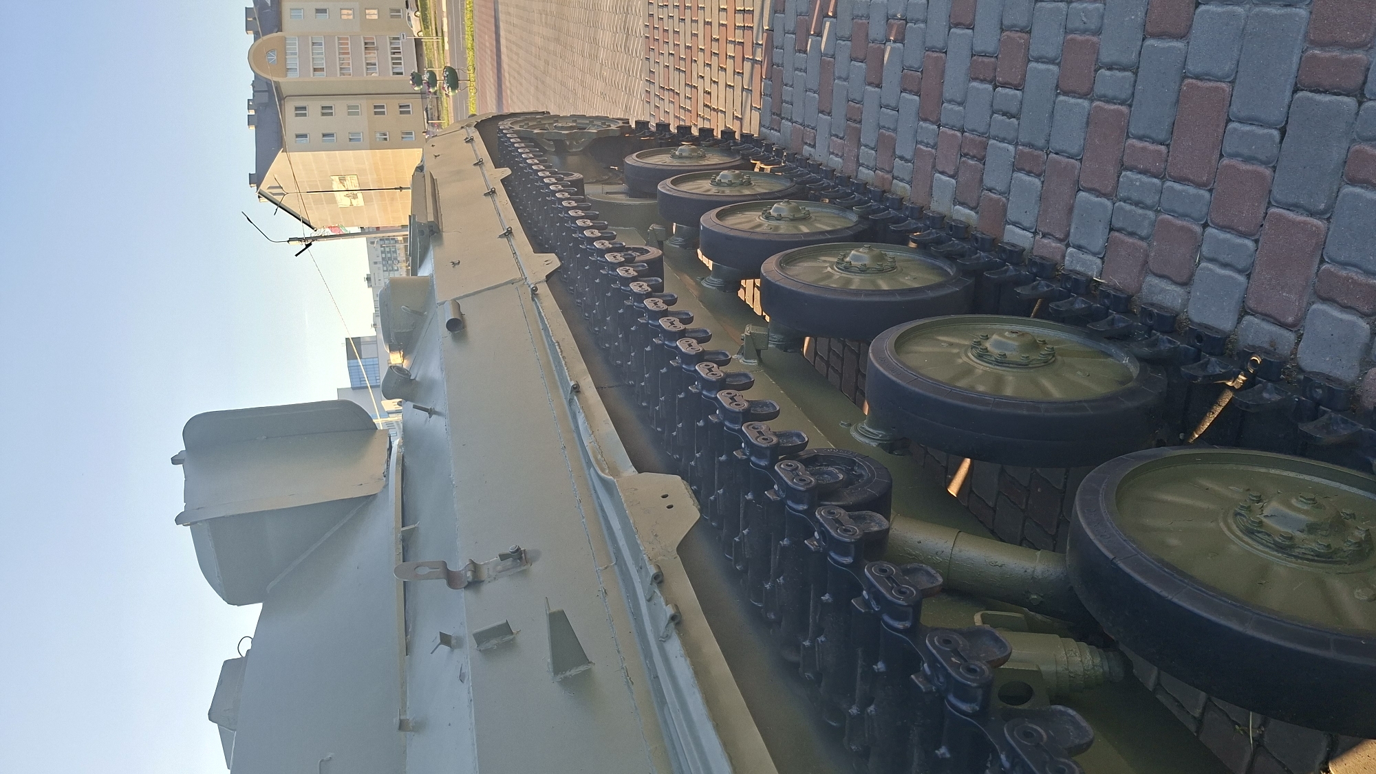 The image captures a close-up, low-angle view of the track and wheels of a military vehicle, likely a tank or armored personnel carrier, on display. The vehicle is painted in a dull green camouflage color and is positioned on a paved walkway constructed of reddish-brown and gray interlocking bricks. The track is made of black metal links with prominent road wheels, some of which are also painted green and appear to have yellow hubs. The scene is illuminated by the warm, low light of what appears to be a sunrise or sunset, casting long shadows and highlighting the texture of the vehicle's tracks and the paving stones.
In the background, a modern, multi-story building with a light-colored facade and numerous windows is visible. This building is situated in the city of Smalyavichy, Belarus. A street with a sidewalk and some greenery is also present in the mid-ground. The sky is clear and a pale blue, indicative of fair weather. The overall impression is of a static display, possibly in a public space or memorial site, given the urban setting and the placement of the military vehicle.