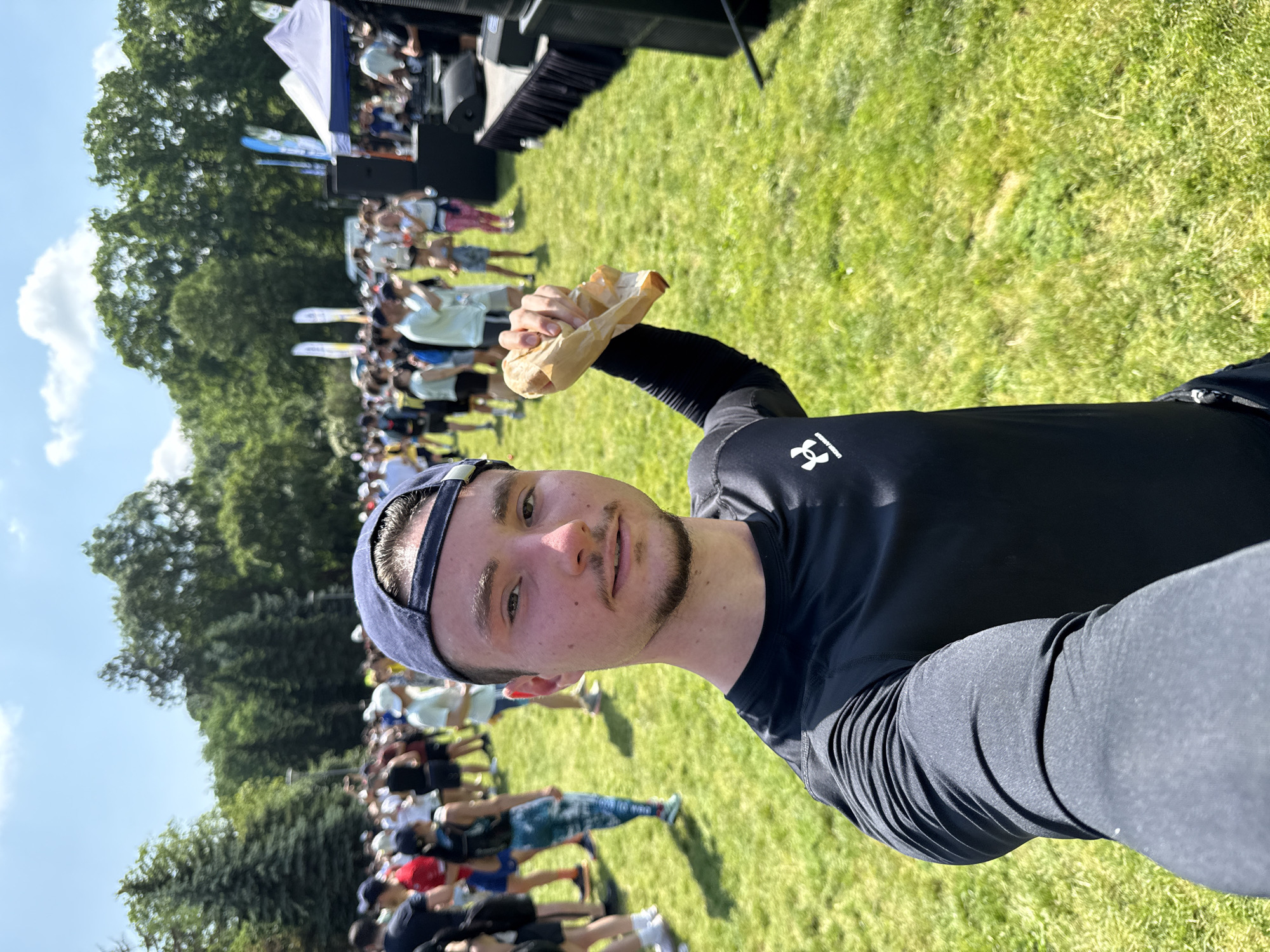A male individual, approximately 20-30 years old, with short brown hair, a short beard, and a mustache, is captured in a selfie at an outdoor event in Sofia, Bulgaria. He is wearing a dark blue baseball cap backward and a black long-sleeved athletic shirt with an Under Armour logo on the left chest. He holds a crumpled light brown paper bag in his raised left hand, angled towards the right side of the frame, and looks directly at the camera with a neutral expression.
The background, heavily blurred, reveals a large gathering of people, many dressed in athletic wear, dispersed across a vibrant green grassy field under a bright blue sky with scattered white clouds. To the right of the frame, a dark-colored stage or platform is visible, equipped with multiple large black speakers. Beyond the stage, white canopy tents and tall white and blue vertical banners are present, indicating an organized event, likely a race or festival. A dense line of mature green trees forms the distant horizon. The specific location is identified as Borisova Gradina Park (Boris' Garden) in Sofia, Bulgaria, a known venue for such public events.