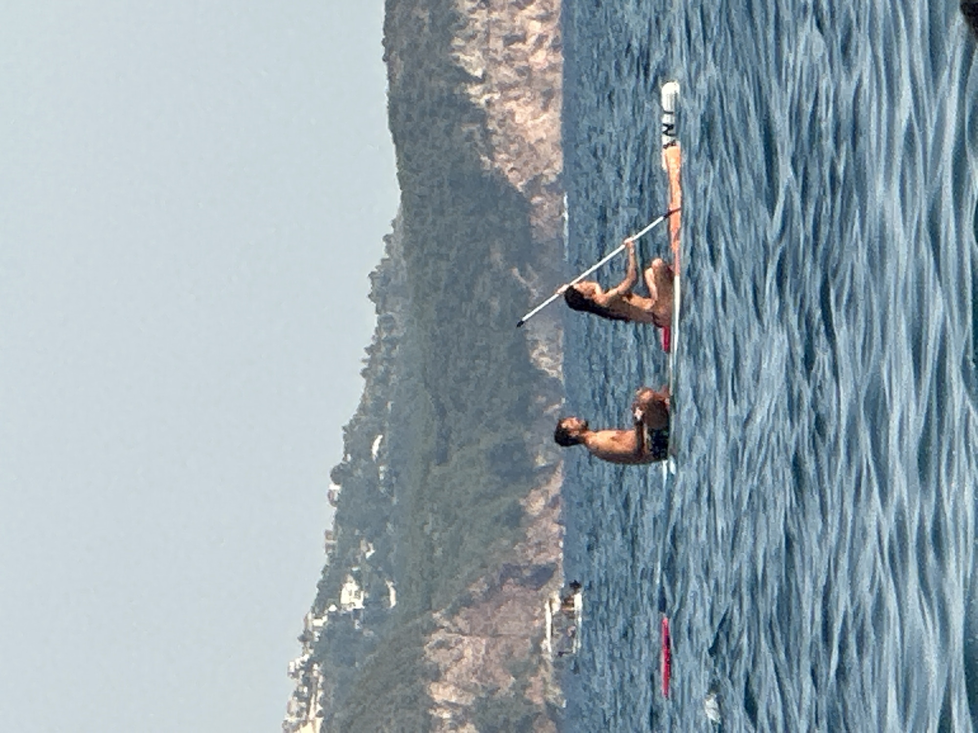 Two people are paddleboarding in the sea. On the left, a man with short dark hair and a beard, wearing dark patterned swim trunks, is seated cross-legged on a paddleboard. On the right, a woman with long dark hair, wearing a bikini, is seated on a paddleboard and holding a paddle. The paddleboard has a white and orange surface with some markings on the tail. In the background, a forested, mountainous coastline with houses is visible, suggesting a location near a populated area. A small boat is seen in the water to the left of the paddleboarders. The water is choppy, indicating a breezy condition. The sky is clear and light blue. The scene is set in the waters off the coast of Boreti, Montenegro, a town known for its beaches and scenic coastline.