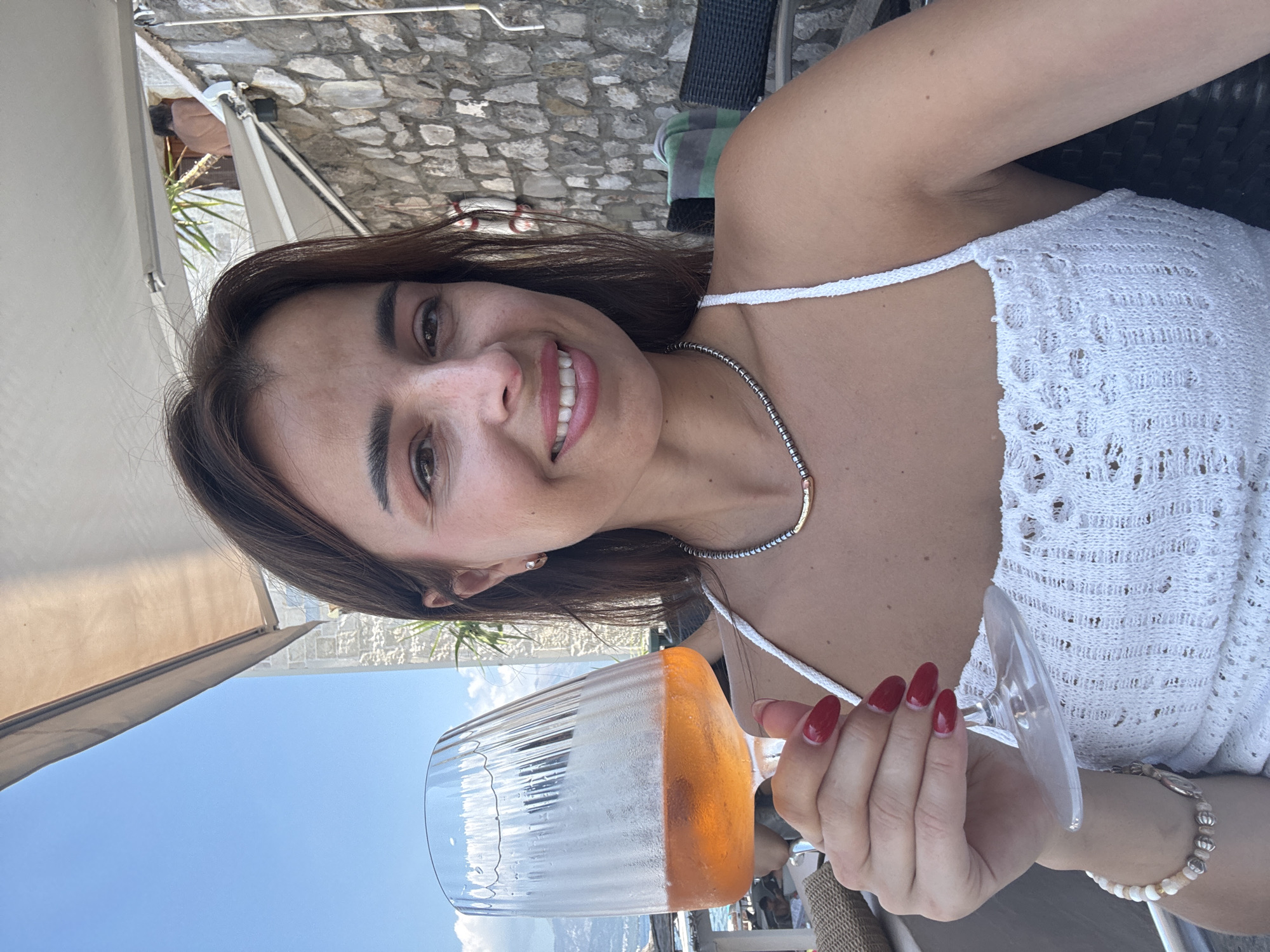 A woman with brown hair smiles as she holds a glass of an orange beverage in a stemmed wine glass. She is wearing a white lace top and a silver necklace. The background shows a stone wall, an awning, and a blue sky.
