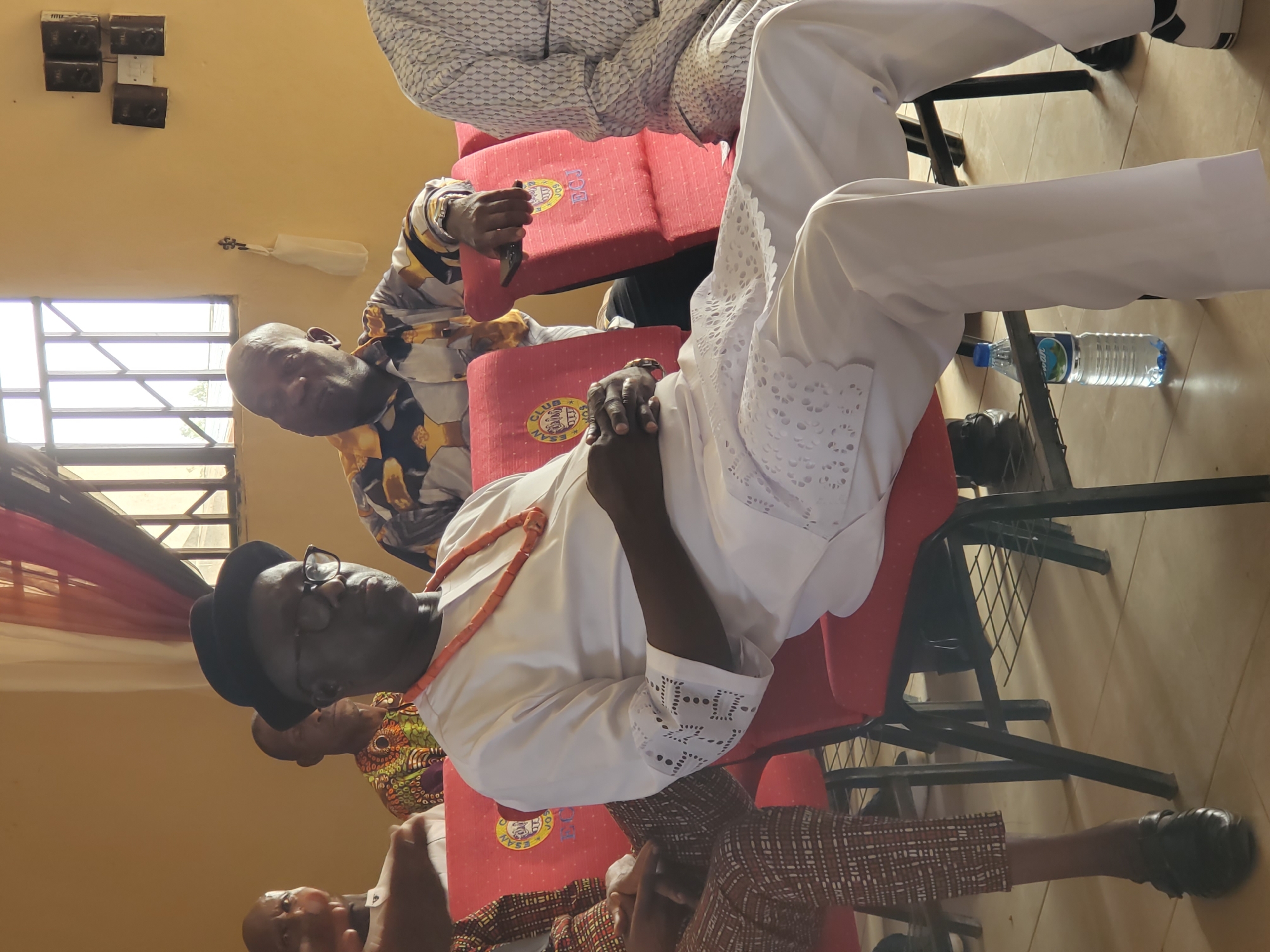The image shows a group of men sitting in red chairs in an indoor setting. One man is wearing a white lace-trimmed outfit with a coral necklace and a black hat and glasses. Another man is wearing a patterned shirt. A water bottle is on the floor near the chairs. The location is likely in the city of Jos, Nigeria.