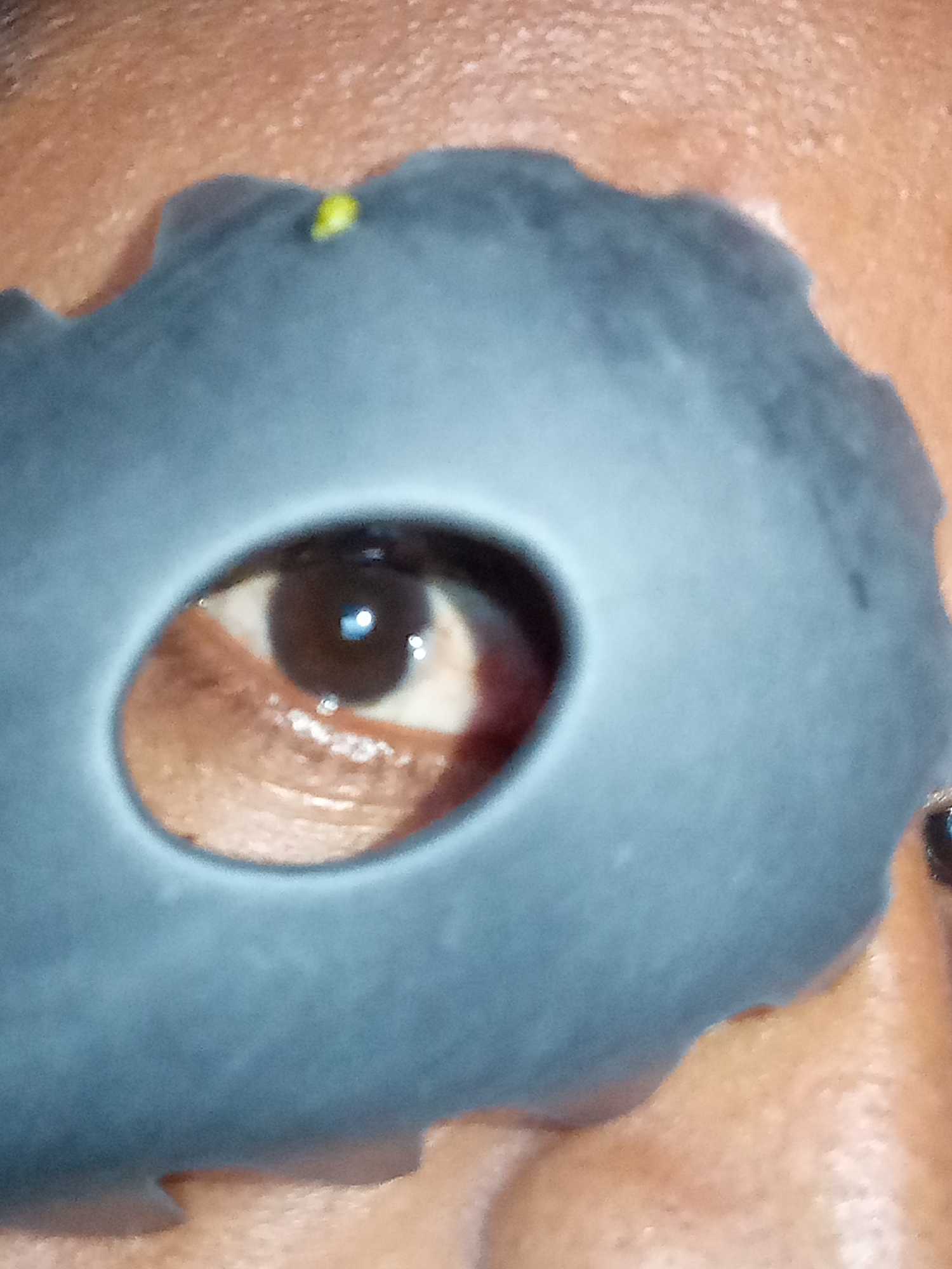 A close-up shot depicts a person's eye visible through a jagged, dark-colored mask with a circular cutout. The skin around the mask appears to be tan. A small, yellow speck is on the mask. The environment is unspecified.