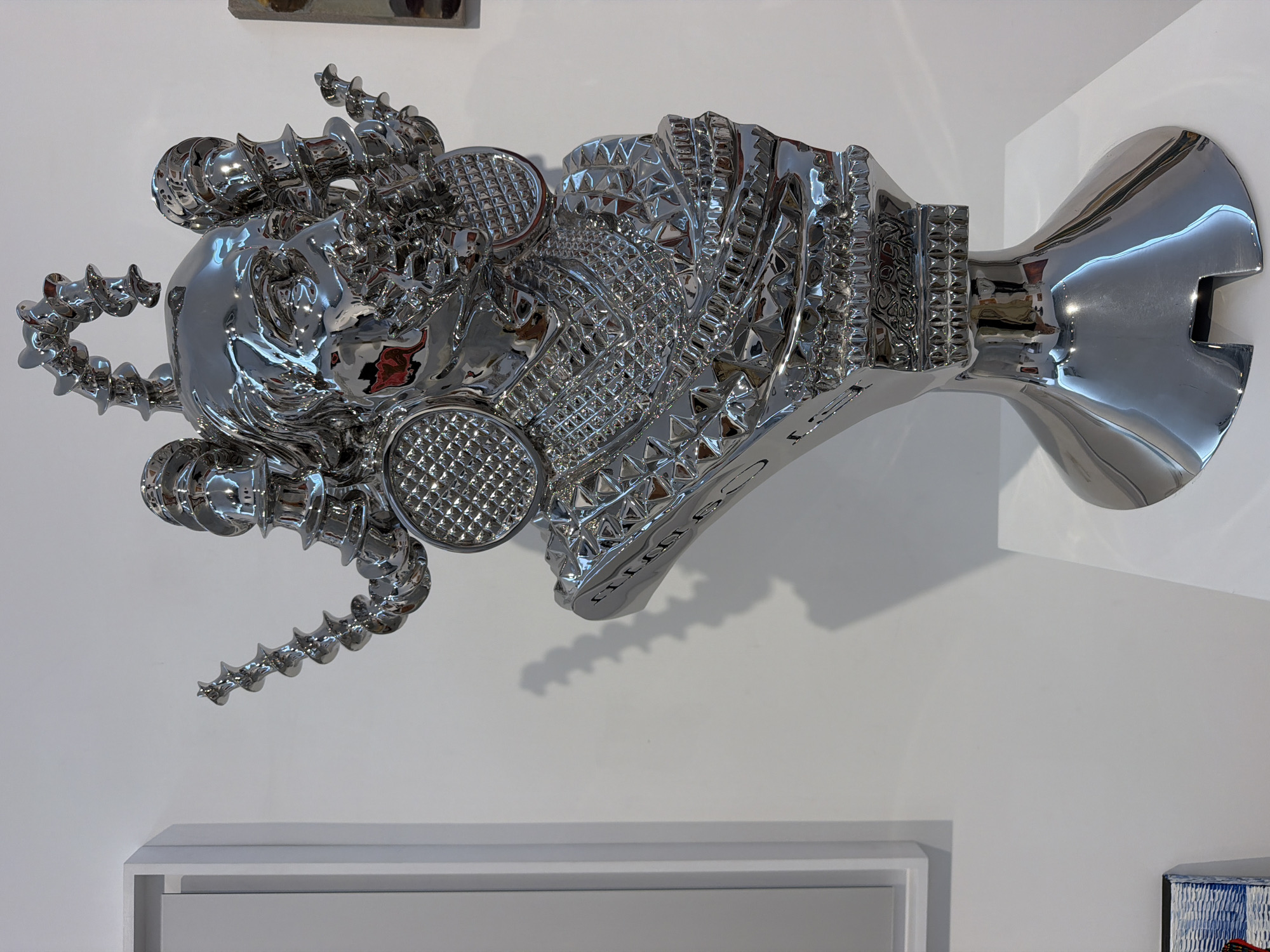 A polished, chrome bust of a figure with an elaborate headdress and face jewelry is displayed in an art gallery. The figure has a serene expression, with its head tilted slightly to the left. The headdress consists of spiraling, screw-like appendages extending upwards and outwards, framing the face. Large, circular earrings with a textured pattern adorn the sides of the head. The bust is adorned with layered, geometric embellishments around the neck and chest, resembling armor or intricate jewelry. The sculpture rests on a flared pedestal, which is placed on a white display surface. The background is a plain white wall, with shadows cast by the sculpture. To the left of the sculpture, a white framed artwork is visible, and a small portion of another framed artwork with a blue and white pattern is visible in the bottom left corner. The lighting in the gallery is bright, creating reflections on the chrome surface of the sculpture. The setting is likely an art exhibition in Tel Aviv, Israel, given the style and presentation of the artwork.