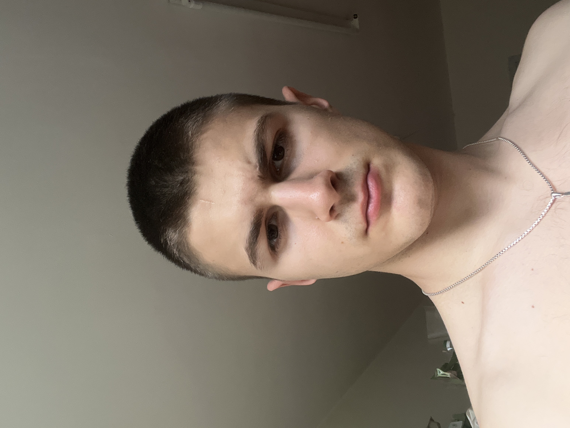A head-and-shoulders portrait of a young man is depicted, with the image rotated 90 degrees clockwise. The man, who appears to be young, has short, dark, buzz-cut hair. He is looking directly at the camera with a neutral to slightly serious expression. His head is tilted towards his right shoulder. A subtle, light-colored scar is visible on his forehead above his left eyebrow. He has fair skin and a faint shadow of stubble around his mouth. He is shirtless, revealing his bare shoulder and collarbone on the right side of the frame, and is wearing a thin silver chain necklace. The background is a plain, light grey wall, indicating an indoor setting. The lighting seems to originate from the left side of the frame, creating a subtle highlight on his left side and a softer shadow on his right. There is no discernible activity taking place other than posing for the portrait. No visible text is present in the image.