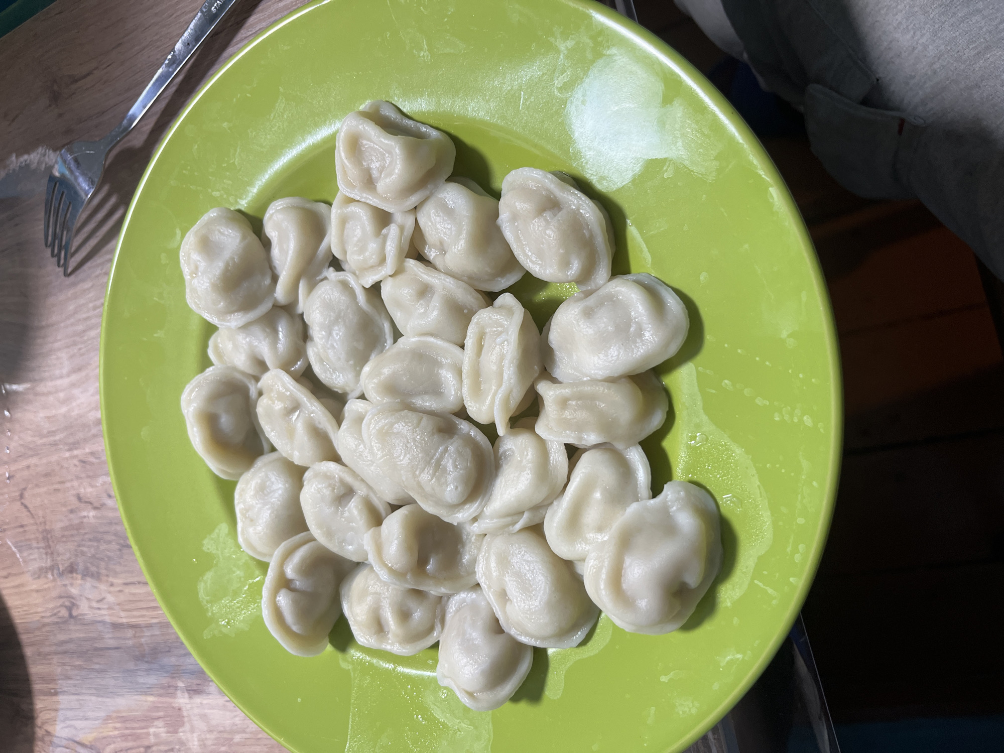 A green plate piled high with boiled pelmeni (small meat dumplings) sits on a wooden surface next to a fork. The pelmeni appear moist, possibly freshly cooked. The location is likely a domestic setting, potentially in Nizhny Tagil, Russia. There are no people clearly visible in the image, although a portion of a gray-colored sleeve can be seen in the upper right corner.