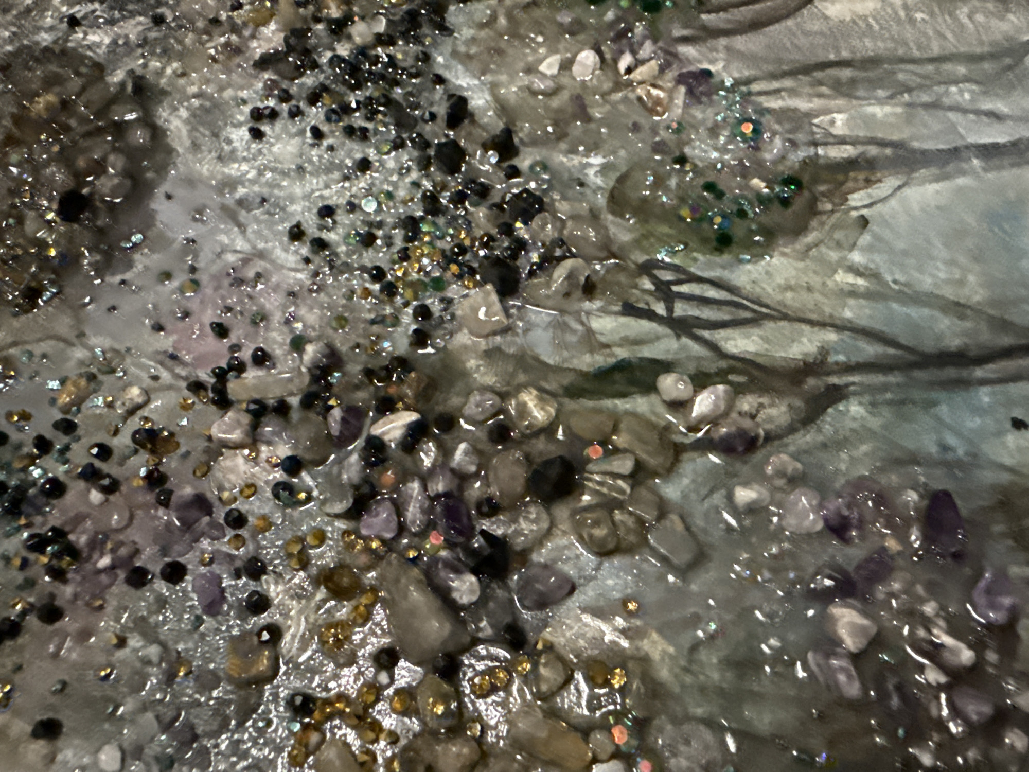 This image, captured in Berezyna, Ukraine, provides a detailed close-up of a mixed-media artwork. The artwork features a highly textured surface composed of numerous small, irregularly shaped stones, predominantly in shades of grey, translucent white, and light purple, embedded within a glossy, resin-like medium that creates a wet, glistening appearance. Scattered throughout this surface are various small, reflective embellishments, including gold-colored beads, dark faceted beads, and iridescent glitter in shades of green, black, and multi-color. In the lower section of the composition, faint, dark, slender lines resembling leafless tree branches are visible against a softly painted background of muted blues, greens, and greys, suggesting a stylized, misty forest or landscape beneath the foreground's textured elements. No people or specific actions are depicted within the scene.