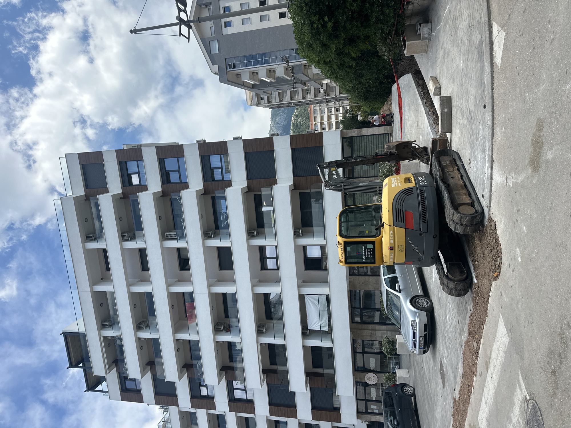 The image captures a street scene in Budva, Montenegro. A compact Volvo excavator is parked on the sidewalk, partially blocking a silver Volkswagen Golf with license plate PG FZ949. The excavator's side displays "T&M commer" and phone numbers. A dark-colored car is parked further down the street.
A modern, multi-story building with a white and wood-paneled facade dominates the left side of the frame. The building features rows of balconies with glass railings and individual air conditioning units. In the background, other buildings and a mountain are visible. The sky is partly cloudy.
The sidewalk appears to be under construction or repair. A section of the pavement has been removed, exposing the earth beneath. Red safety tape is visible in the background. Two people can be seen in the distance near the exposed earth.