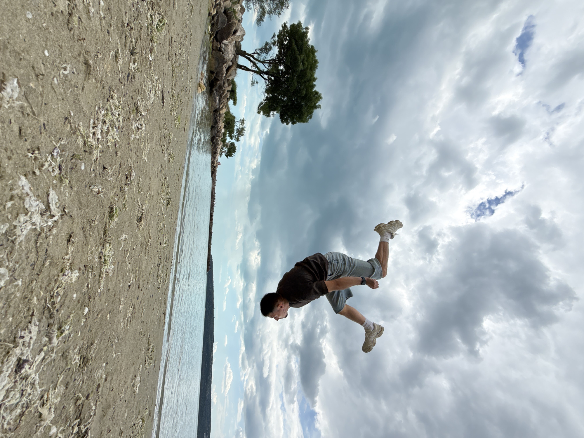 A man is in mid-air, seemingly performing a stunt, with his body parallel to the ground. He is wearing a dark-colored shirt, light-colored shorts, white socks, and running shoes. The background is composed of a beach, water, and a cloudy sky. A tree can be seen at the horizon. The location is likely a beach in Varna, Bulgaria.
