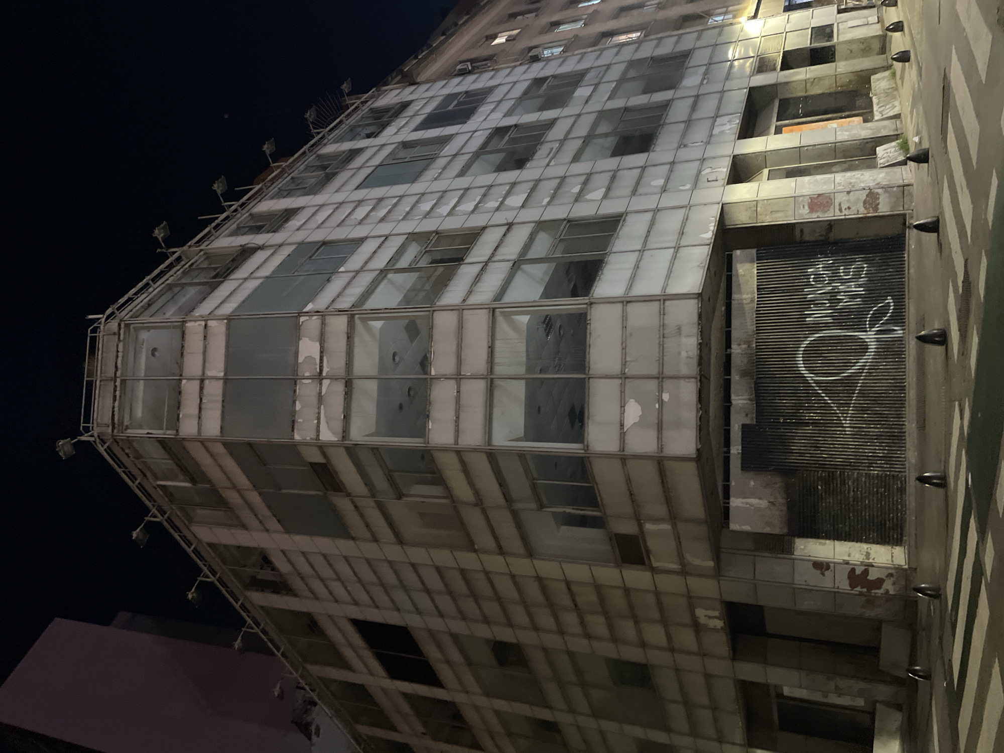 The image shows a tall, deteriorating building in Buenos Aires, Argentina. The building has a grid-like facade with many windows, some of which are broken or missing. The building's lower floors have a closed-off section with graffiti. The sky is dark, and streetlights are visible at the top of the building. The building appears abandoned or neglected.