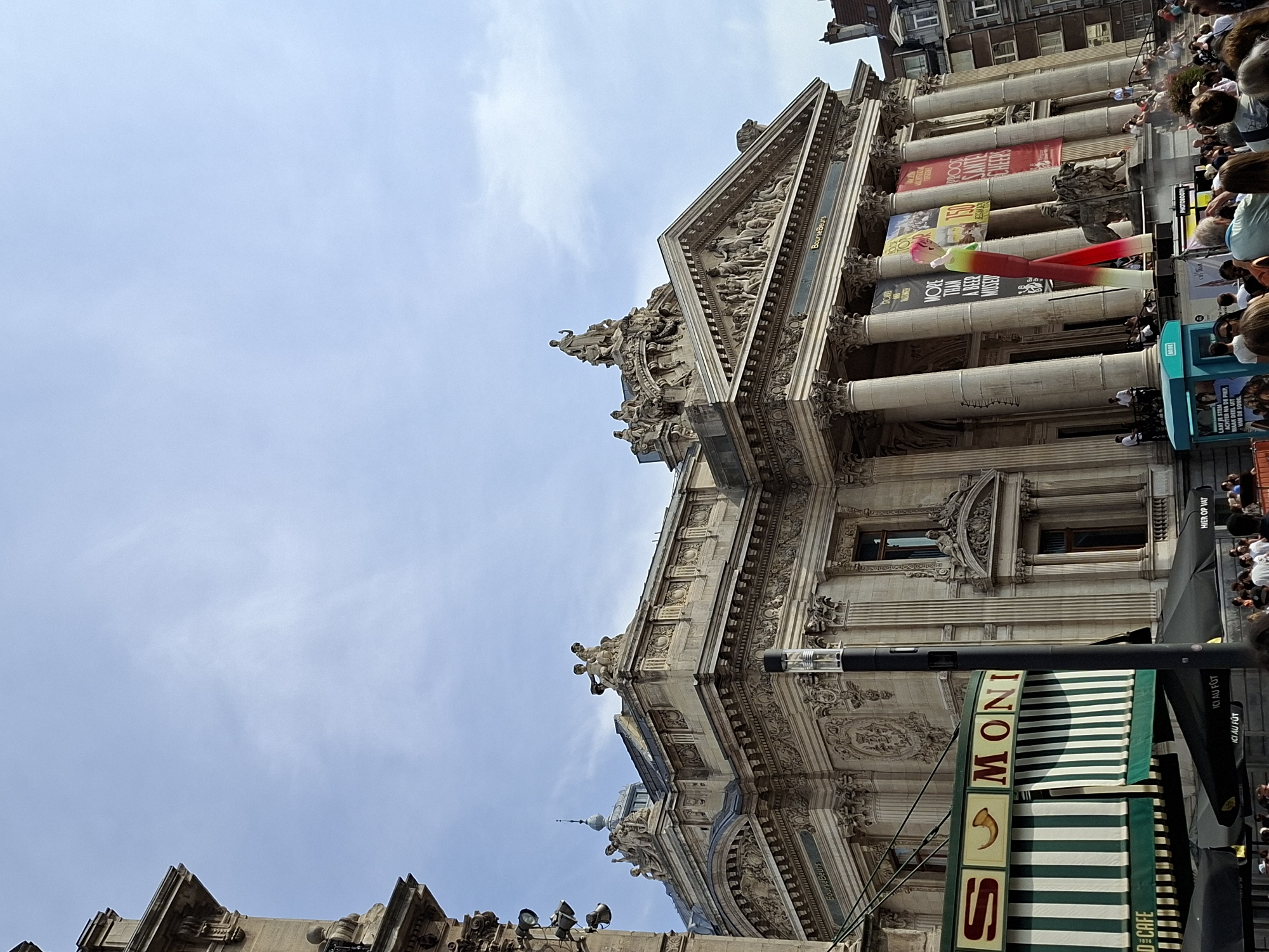 This image depicts a street view in Brussels, Belgium, featuring the exterior of the Royal Stock Exchange building. The grand, ornate architecture of the building is prominent, with classical columns, intricate carvings, and statues adorning its facade. In front of the building, a tall, colorful inflatable figure stands, appearing to be a whimsical art installation. Several banners are displayed on the building, some featuring text and images related to events or exhibitions. In the foreground, a crowd of people is gathered, some seated at outdoor tables under umbrellas, suggesting a lively urban atmosphere. The sky above is partly cloudy. A cafe with green and white striped awnings is visible to the left, bearing the name "MONI" and "O DE CAFE". A dark blue kiosk with text and images is also present in the lower left portion of the frame. The overall scene suggests a public space in a bustling European city.