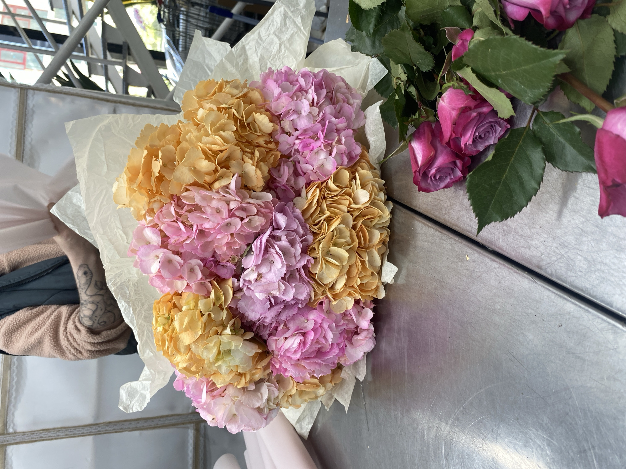 Here is a detailed description of the image:
The image shows a close-up, top-down view of a bouquet of hydrangeas and roses. The hydrangeas are a mix of orange and pink hues, arranged in a cluster and wrapped in white tissue paper. To the right of the hydrangeas lie several pink roses with green leaves. These flowers rest on what appears to be a metallic surface, likely a counter or table.
In the lower left corner of the frame, a portion of a person's arm is visible, clad in a beige and green jacket. The arm also features a tattoo.
The scene appears to be indoors, possibly in a florist shop or similar setting. It is not possible to definitively confirm the location as Yekaterinburg, Russia based on the information present in the image.