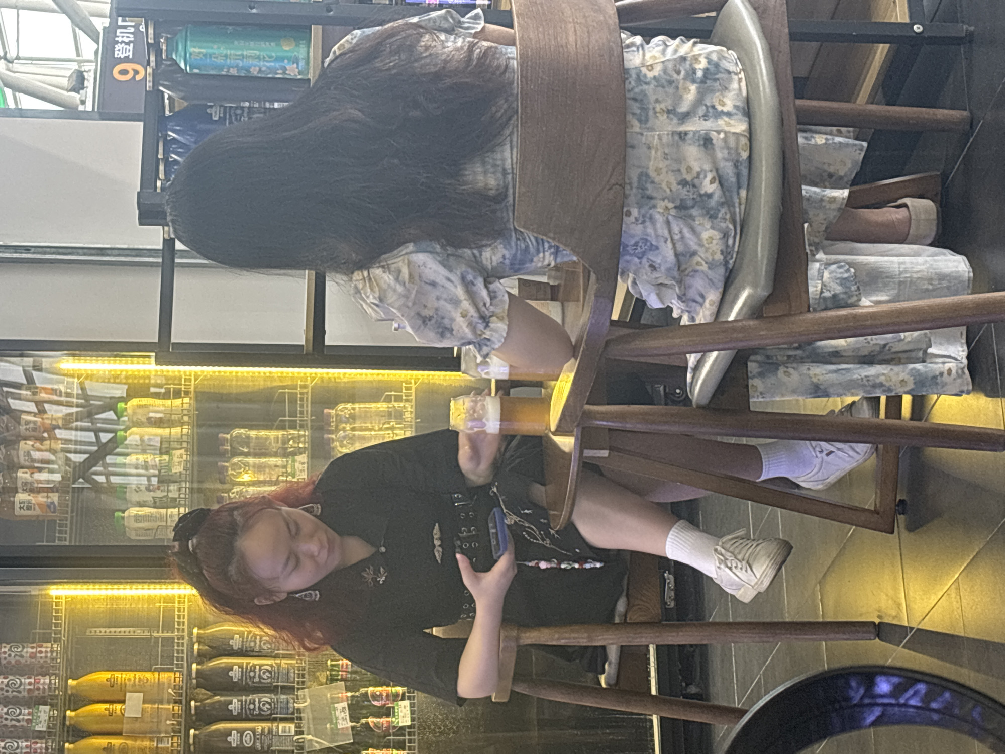 Two women are seated at tables in a dimly lit establishment, possibly a bar or cafe, with a refrigerated display case filled with beverages visible behind them. The woman on the left, with bright red hair, wears a dark outfit with a belt and large earrings. She is holding a dark smartphone and looking down. A glass of amber liquid, likely beer, sits on the table in front of her. The woman on the right has long black hair and wears a floral print dress. She is facing away from the camera, and her arms are resting on the table. The environment appears to be indoors, with a focus on the two individuals and the products in the display case. The scene is set within Xinwu District, China.