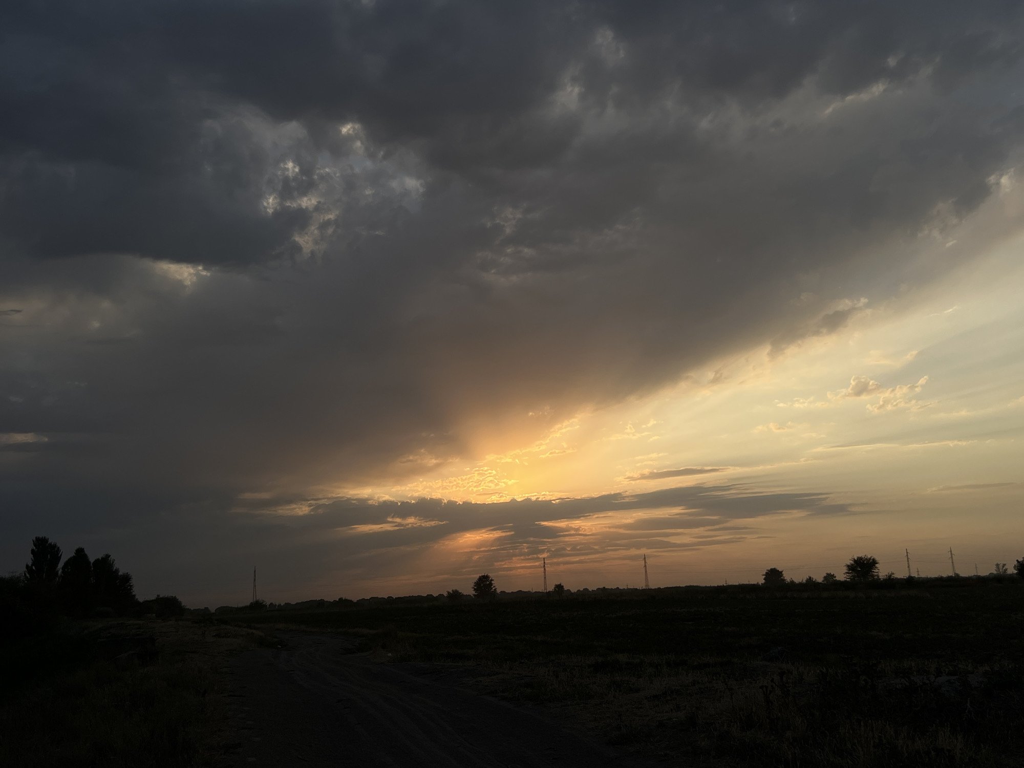 The image depicts a rural landscape at dusk, with a dramatic sky filled with dark, heavy clouds interspersed with areas of golden light. The sun is setting, casting a warm glow through a break in the clouds, illuminating the horizon with hues of orange and yellow. The foreground features a dark, unpaved road or path leading into the distance, bordered by silhouetted vegetation and trees on the left side. In the middle ground and background, the land appears flat and dark, with sparse trees and utility poles visible against the twilight sky. The overall atmosphere is somber and dramatic due to the stormy sky and low light conditions. The location is described as being in the city of Drujba, Kyrgyzstan.