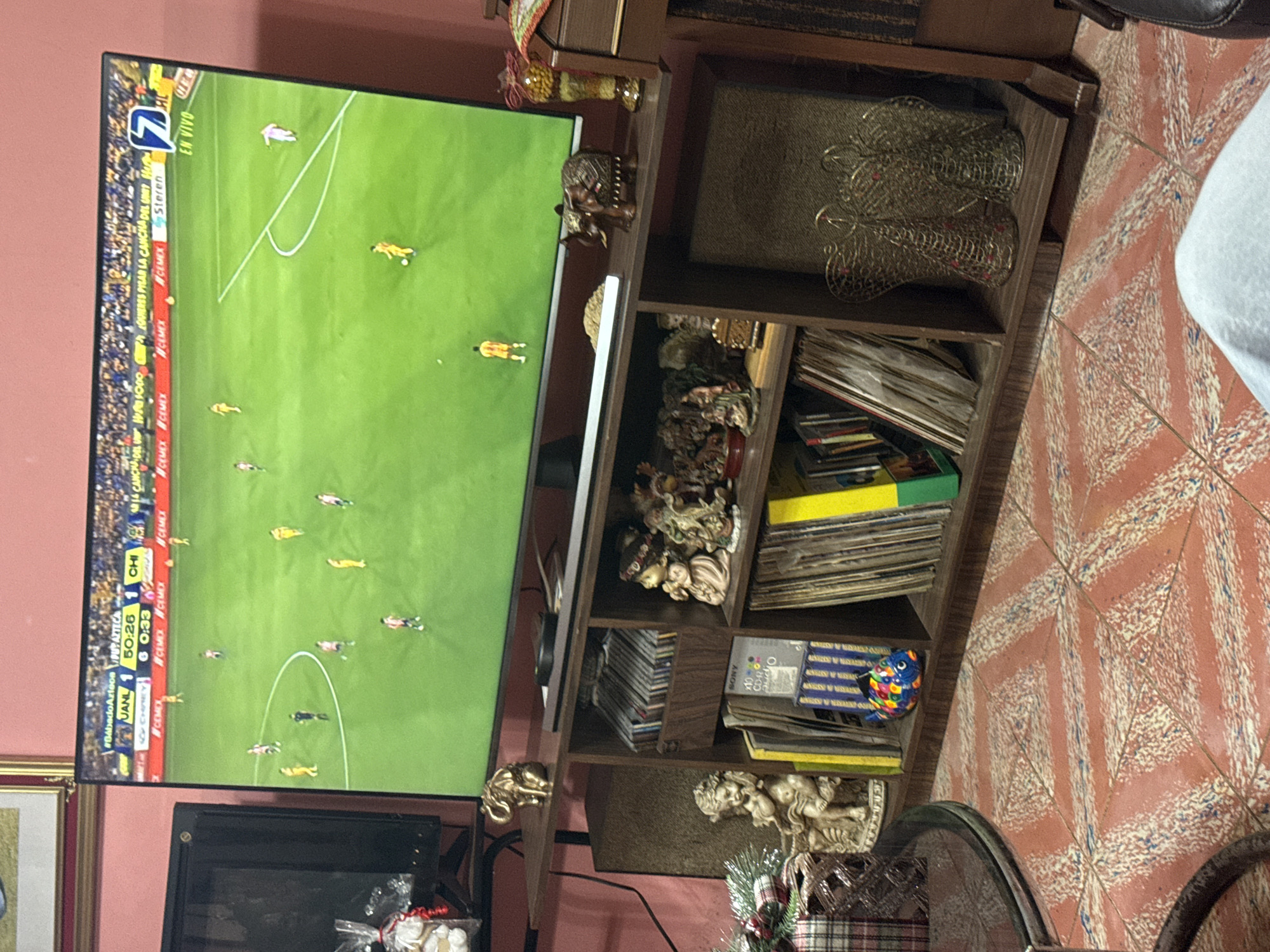 The image shows a television displaying a soccer game, with the score indicating "JANL 1" and "CHI 6" at the 50:26 mark of the first half. The TV is located in a room with a patterned tile floor and a brown wooden shelf filled with items, including decorative figures, CDs, and other objects. The overall environment suggests a residential setting. There are no explicit indications or recognizable landmarks to definitively place the location in San Cristóbal, Mexico; this location is inferred only based on details from the TV and OCR text. There are no people physically present in the frame.