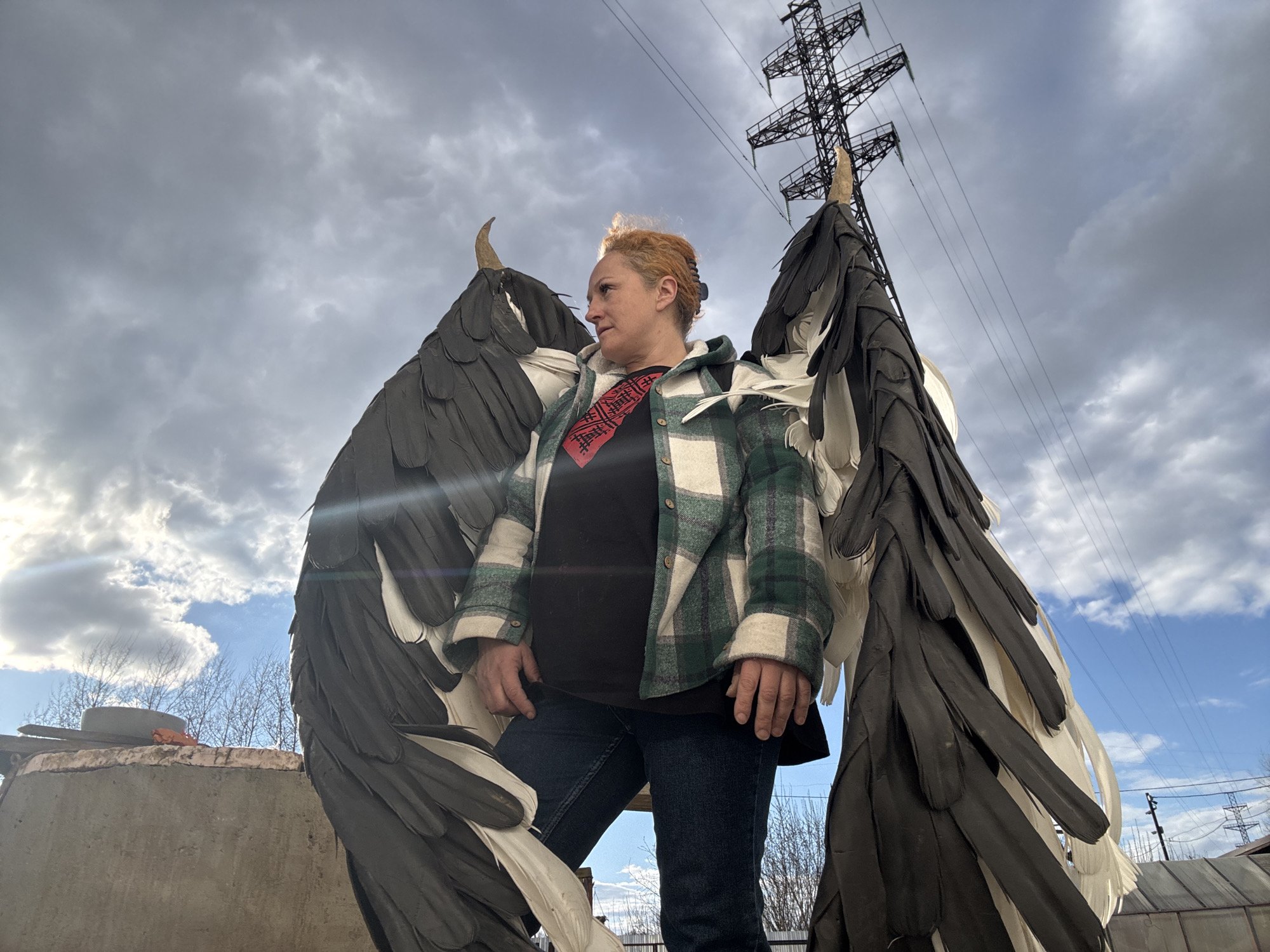 The image depicts a woman outdoors in Chaikovsky, Russia, wearing large, elaborate black and white artificial wings. She has reddish-blonde hair pulled back and is dressed in a green and white plaid jacket over a black t-shirt and blue jeans. A red scarf or tie with an intricate geometric pattern is visible around her neck. She stands with her right foot slightly elevated, looking contemplatively towards her left (viewer's right).
The scene is set under a dramatic sky, characterized by thick grey clouds, though patches of blue sky and sunlight are visible, particularly on the left side. A prominent black metal electricity transmission tower dominates the upper background, silhouetted against the sky. Bare trees are discernible in the distance. The woman appears to be standing on a rough concrete structure. The large wings suggest a costume, art installation, or a theatrical element. The overall atmosphere implies a cool season, such as late autumn or early spring, given the bare trees. No legible text is visible in the image.