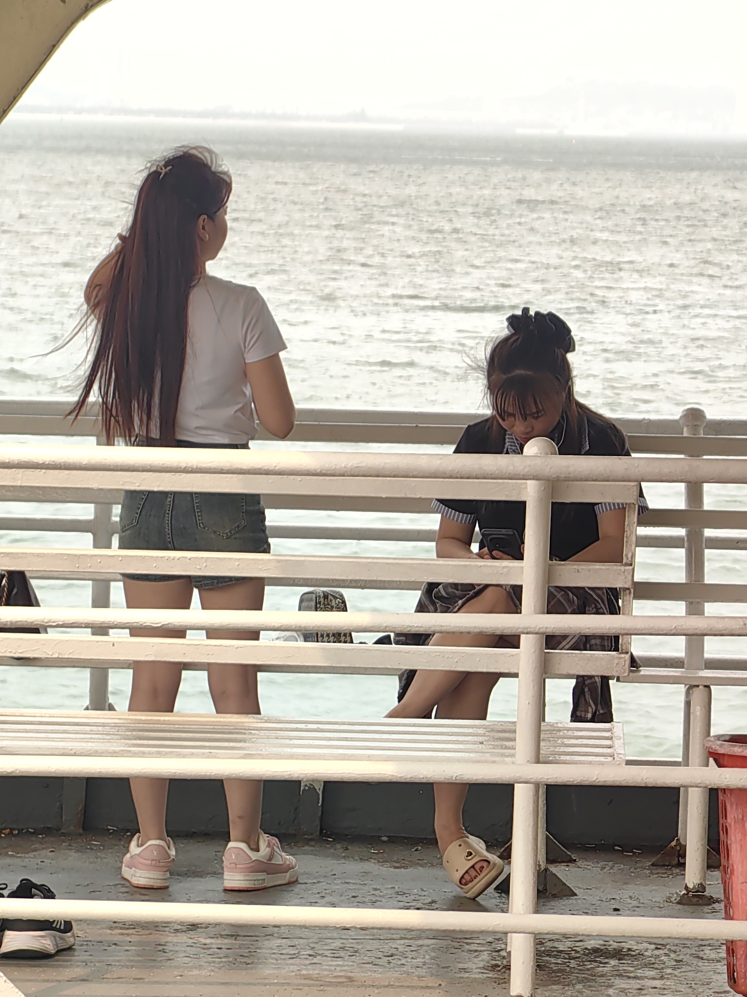 Two young women are on a boat in Tuan Chau Ward, Vietnam. One woman stands facing the ocean, wearing a white shirt and denim shorts. The other woman sits on a white bench looking at a smartphone. The background shows a greyish body of water and hazy skyline. A white railing encloses the area.