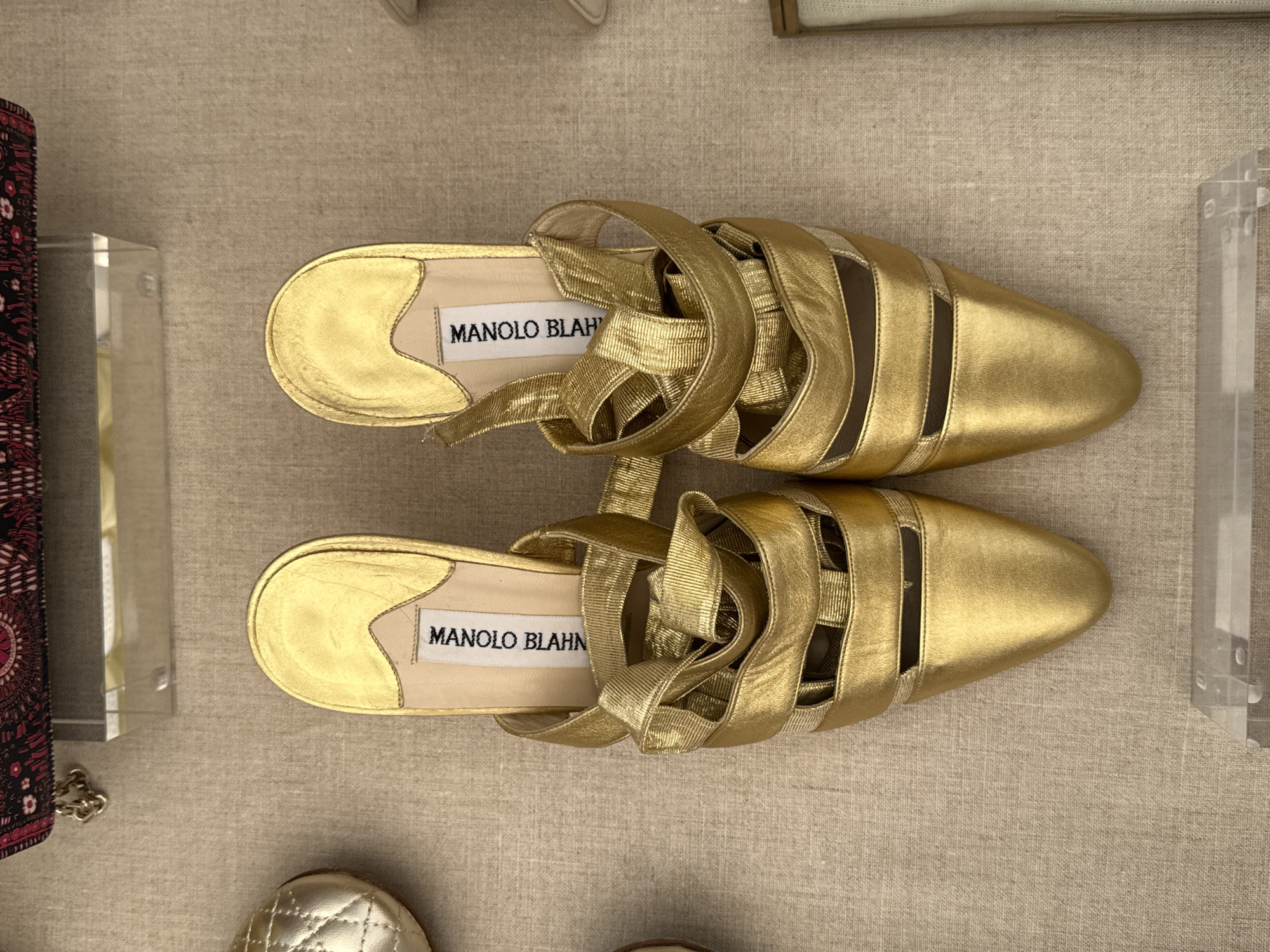 A pair of gold Manolo Blahnik mule shoes are displayed on a tan linen surface, possibly in a retail setting or exhibition space in Mexico City, Mexico. The shoes are angled slightly away from each other. The Manolo Blahnik brand name is clearly visible on the insole of each shoe. Additional objects are partially visible at the borders of the frame, including a decorative item to the left and a transparent object to the right. A portion of another shoe is visible near the bottom left corner.