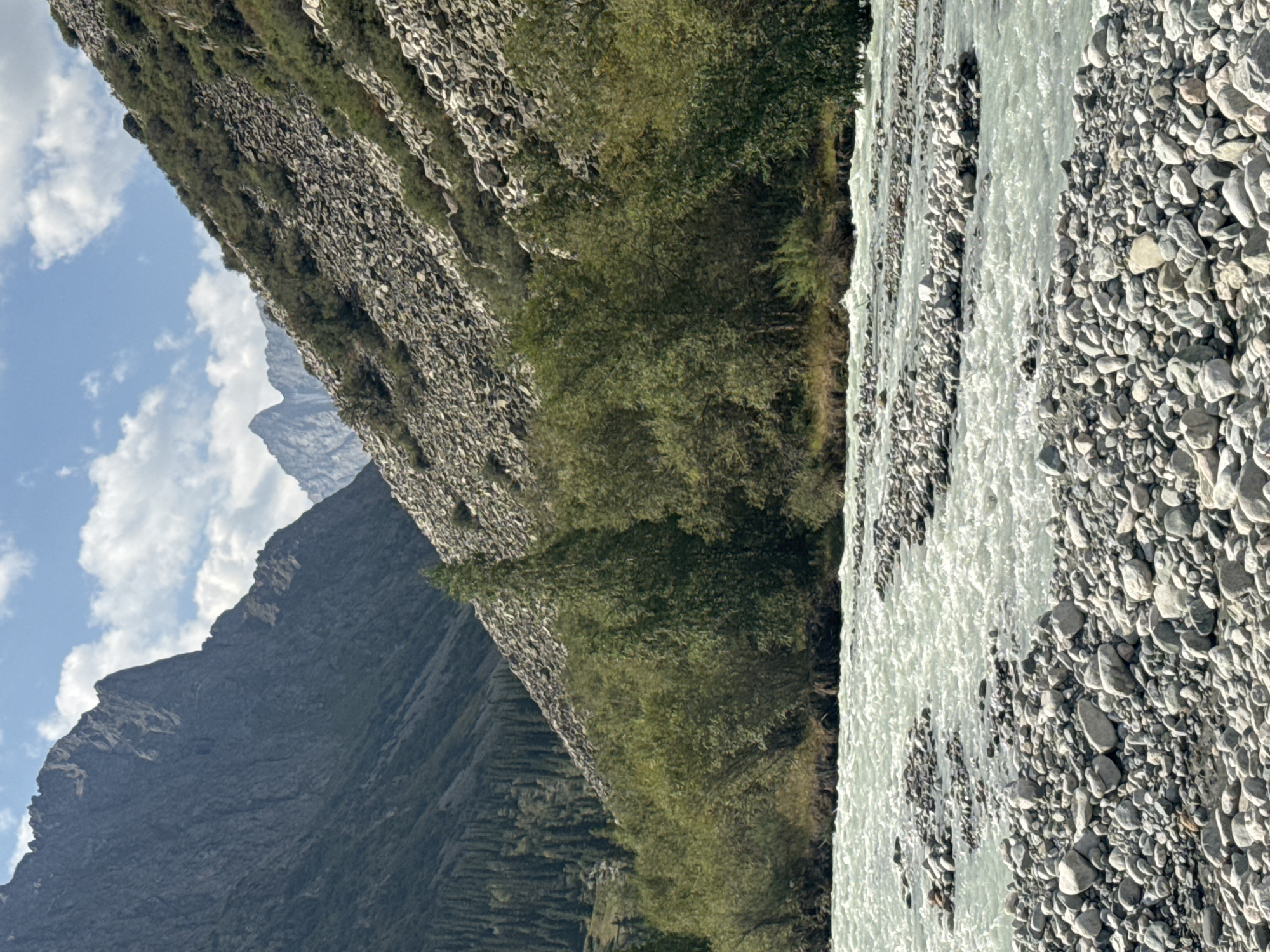 An eye-level, medium shot captures a wide, shallow, stony river flowing through a valley in Kyrgyzstan. The river occupies the foreground and midground, its water a milky turquoise, disturbed by ripples and broken by several gravel bars. The riverbanks are lined with an abundance of grey and white rocks of various sizes, some appearing to be partially submerged. On the far bank, a dense collection of green trees and shrubs grows. Beyond the trees, steep, rocky hillsides rise sharply, covered with a scree of grey stones and sparse green vegetation. In the background, tall, majestic mountains with sharp, jagged peaks pierce the blue sky, which is dotted with fluffy white clouds. The overall scene depicts a rugged, natural landscape.