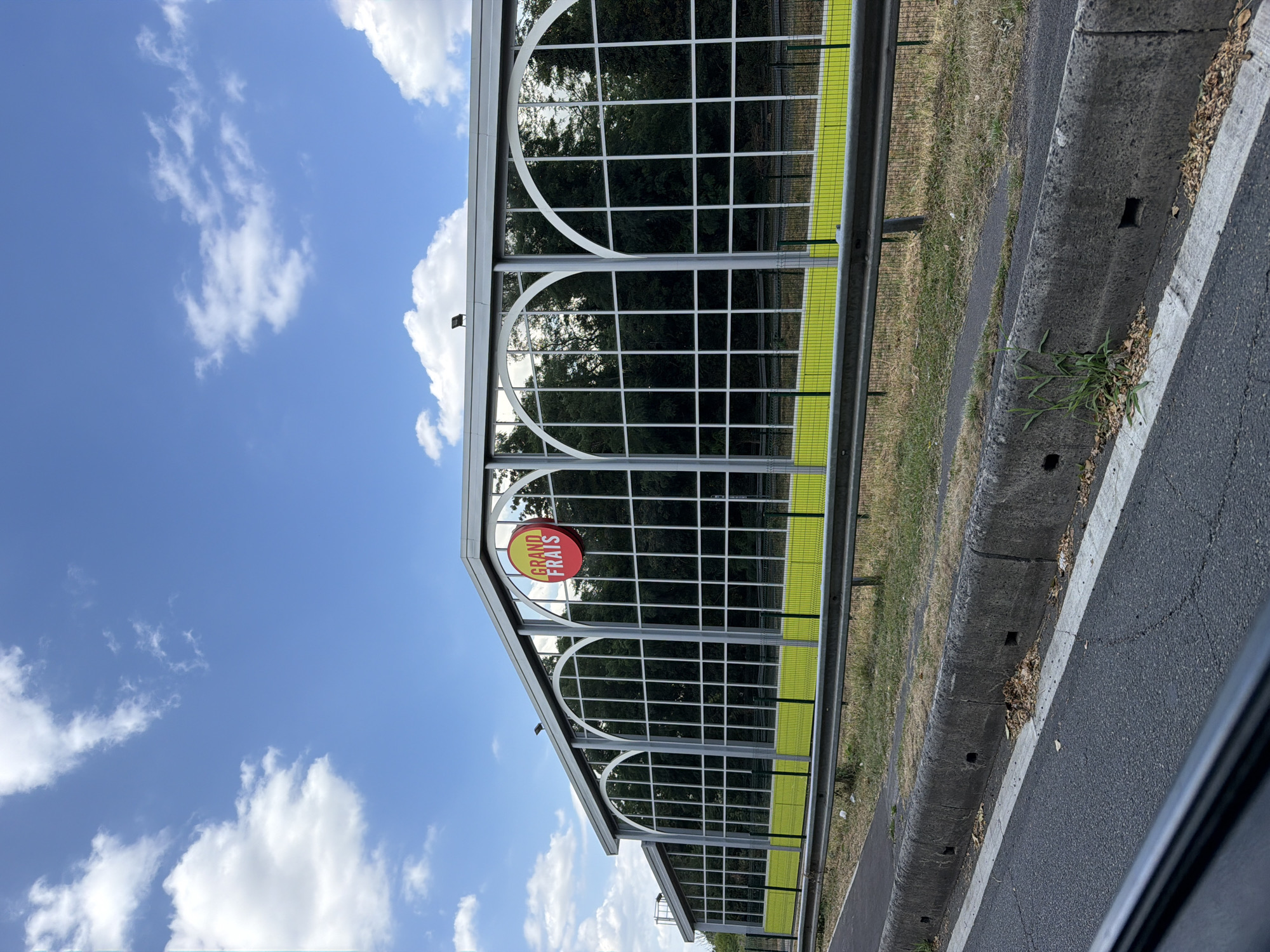 This media file shows a roadside view under a bright, partly cloudy sky. The scene is dominated by a modern, tall sound barrier, likely a noise wall, constructed with metal framing and large glass panels. The glass reflects the surrounding trees and the blue sky with scattered white clouds, suggesting it's daytime. The sound barrier has a distinctive design with a series of arched sections at the top, each filled with a grid of square panes. A bright yellow-green trim runs horizontally along the lower portion of the barrier.
A circular red sign with yellow text is prominently displayed on one of the glass panels, partially obscured by the structure's frame. The text on the sign is "GRAND FRAS".
To the right of the sound barrier is a road surface with a white line marking. A concrete curb separates the road from a strip of dry grass and weeds. The overall impression is of a highway or a major road bordered by this protective barrier. The lighting suggests it is likely mid-morning or afternoon. There are no people visible in the image. The setting appears to be an urban or suburban environment in Évry-Courcouronnes, France, given the location context.
