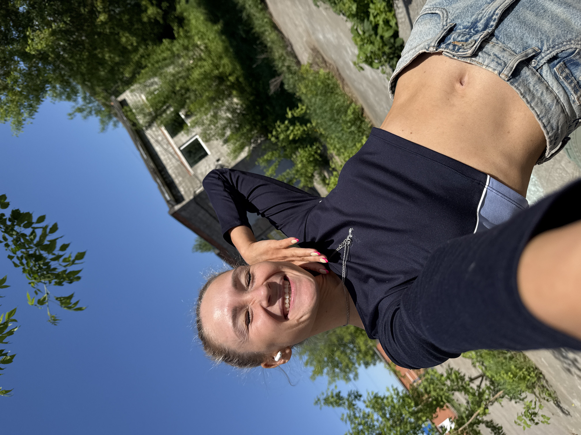 A low-angle, rotated, brightly-lit selfie shows a smiling white woman in a navy blue long-sleeved top and blue denim shorts, standing outdoors. She is facing the camera with her hand near her head, and a white wireless earbud is visible in her left ear. Behind her is a derelict two-story building with trees and vegetation. The background includes a clear blue sky and more trees. The photo was taken outside in Chaikovsky, Russia.