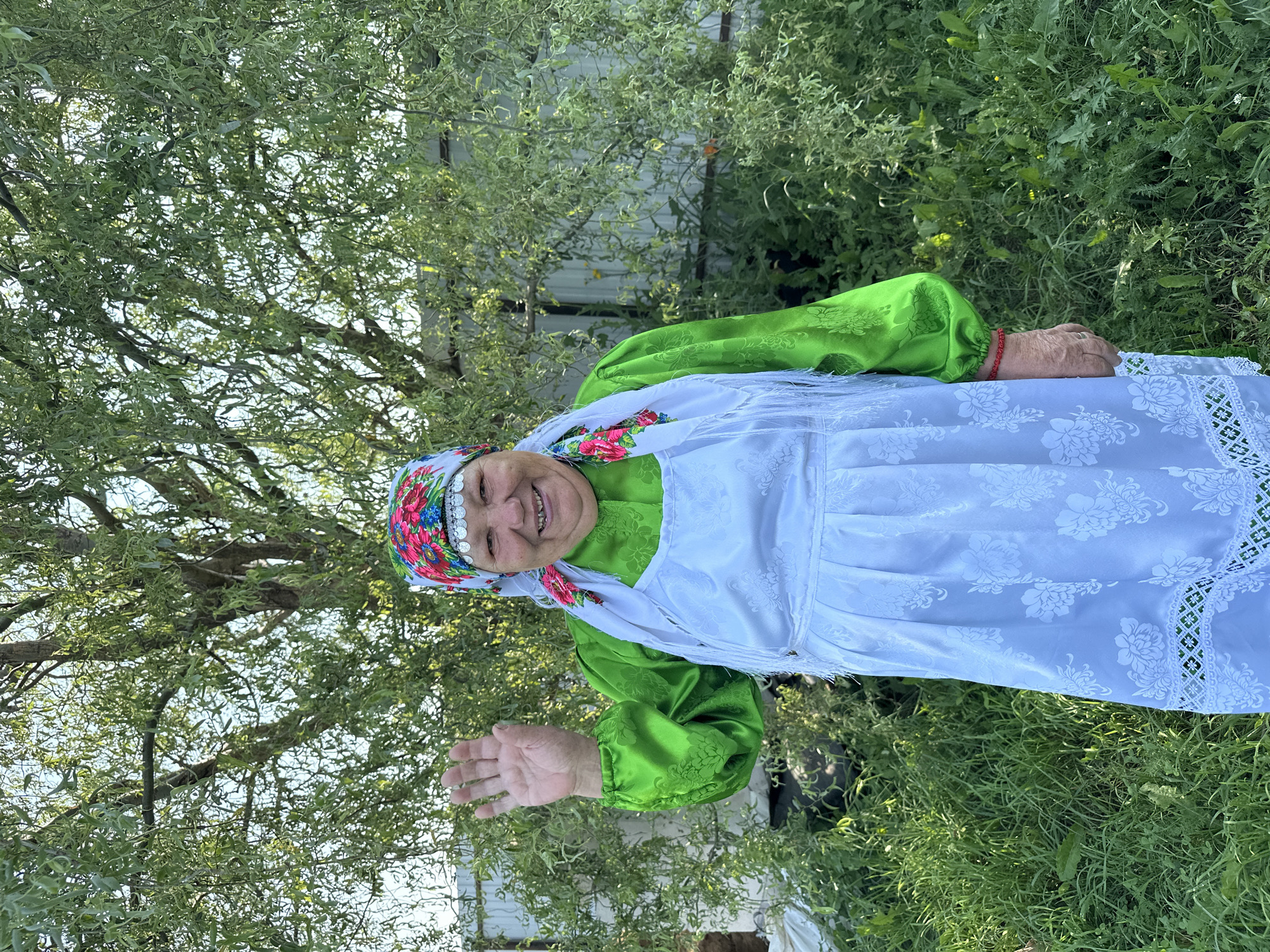 A middle-aged woman stands outdoors, in front of a large tree and a fence. She is wearing a brightly colored headscarf with floral patterns, a green blouse with puffed sleeves, and a white apron with embroidered floral designs. The apron has a fringed edge. Her left hand is raised, palm facing outward, as if waving. She is smiling. The background consists of lush green foliage, tall grass, and a light-colored fence. The lighting suggests it is daytime, likely late afternoon, with the sun casting dappled shadows through the leaves. The location is specified as Sredneye Kechevo, Russia.