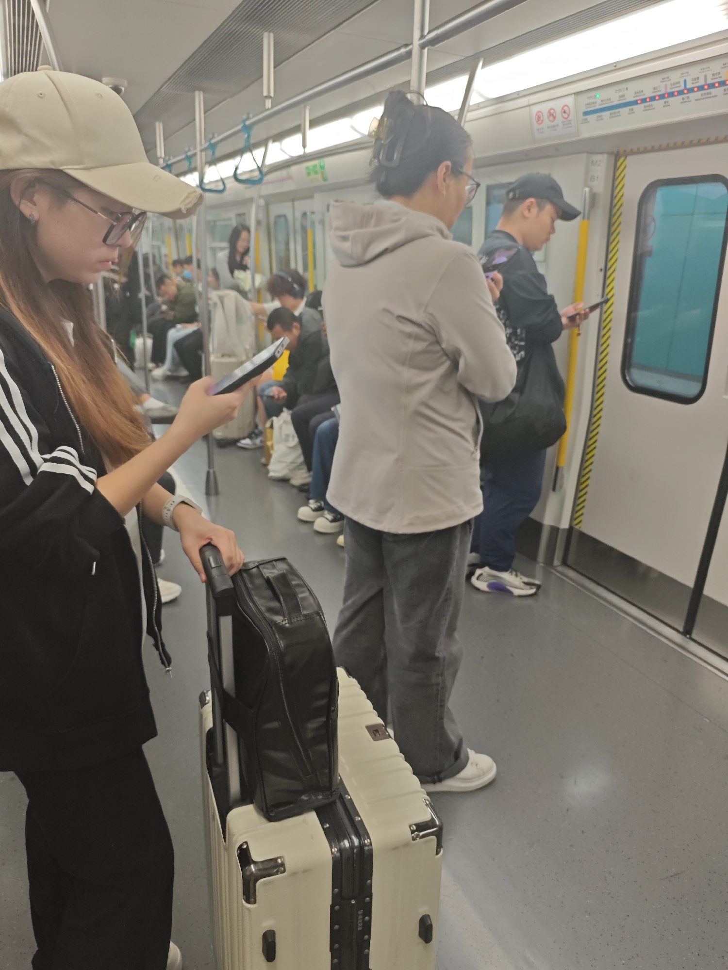 Here is a detailed description of the image:
The image shows a group of people riding on a subway car. The car appears to be in Unknown, Unknown.
In the foreground on the left, a young woman with long brown hair and glasses is wearing a light-colored baseball cap, a black jacket with white stripes on the sleeves, and black pants. She is looking down at her smartphone in her hand. Next to her is a suitcase. The suitcase is cream-colored with black trim. A black travel bag is sitting on top of the suitcase.
Next to the woman is another person. This person is wearing a light gray sweatshirt and dark gray pants. This person is also looking at a phone.
Next to this person is a man wearing a black baseball cap and black clothing. He is also looking at a phone.
In the background, there are other people sitting and standing in the subway car. The subway car has metal handrails and handles. The windows are lined with route maps and informational signs. The doors are light gray with a vertical yellow stripe next to them.