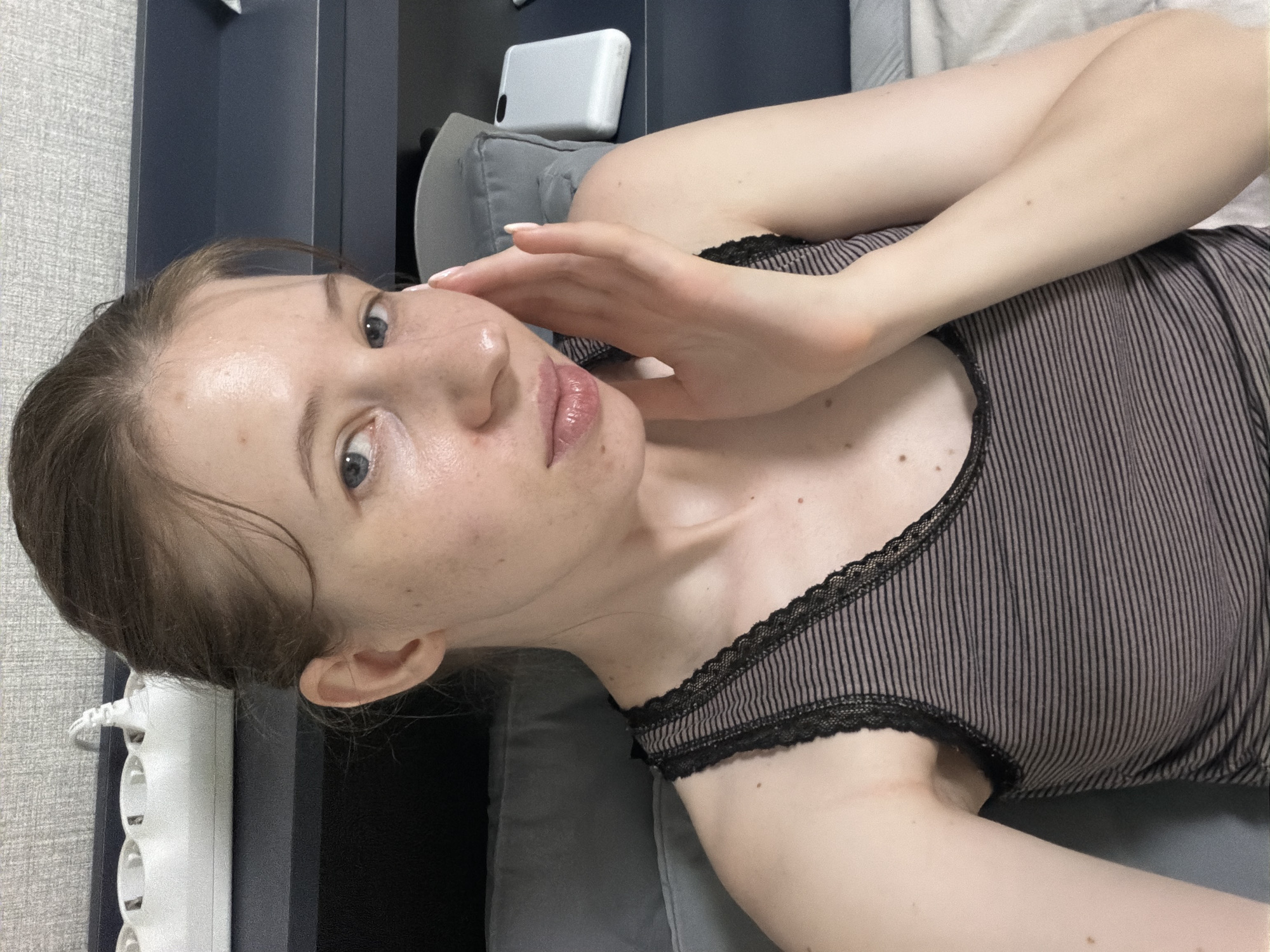 An over-the-shoulder shot shows a young woman with light skin and freckles, her hair pulled back. She is wearing a striped tank top with lace trim and is touching her cheek with her right hand. Her eyes are blue, and her lips are slightly parted. Behind her, a white power strip with multiple outlets is visible against a textured gray background. Further back, a dark blue shelf is present, with a white object, possibly a phone, resting on it. The woman is lying on a bed with gray pillows, and a portion of a white sheet is visible. The scene appears to be in a bedroom, potentially in Dongducheon-si, South Korea.