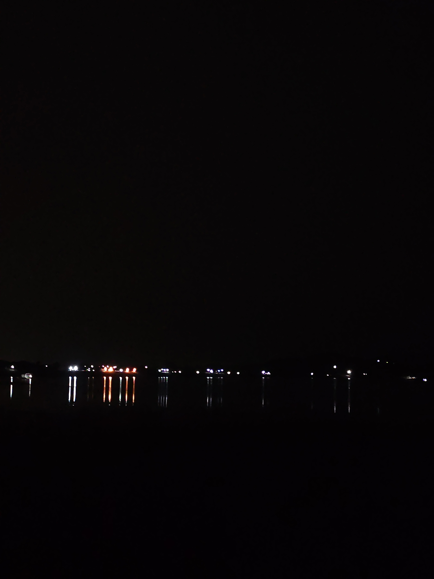 The image depicts a nighttime scene of a body of water, likely a river or lake, with a distant shoreline illuminated by various lights. The lights appear to be from buildings, streetlights, or possibly boats, and their reflections are visible on the dark water's surface, creating elongated streaks of light. The overall atmosphere is dark and quiet, characteristic of a nocturnal setting. The image's resolution and lighting conditions suggest it was taken with a low-light camera setting. No people or specific landmarks are clearly identifiable, but the presence of lights indicates a populated area.