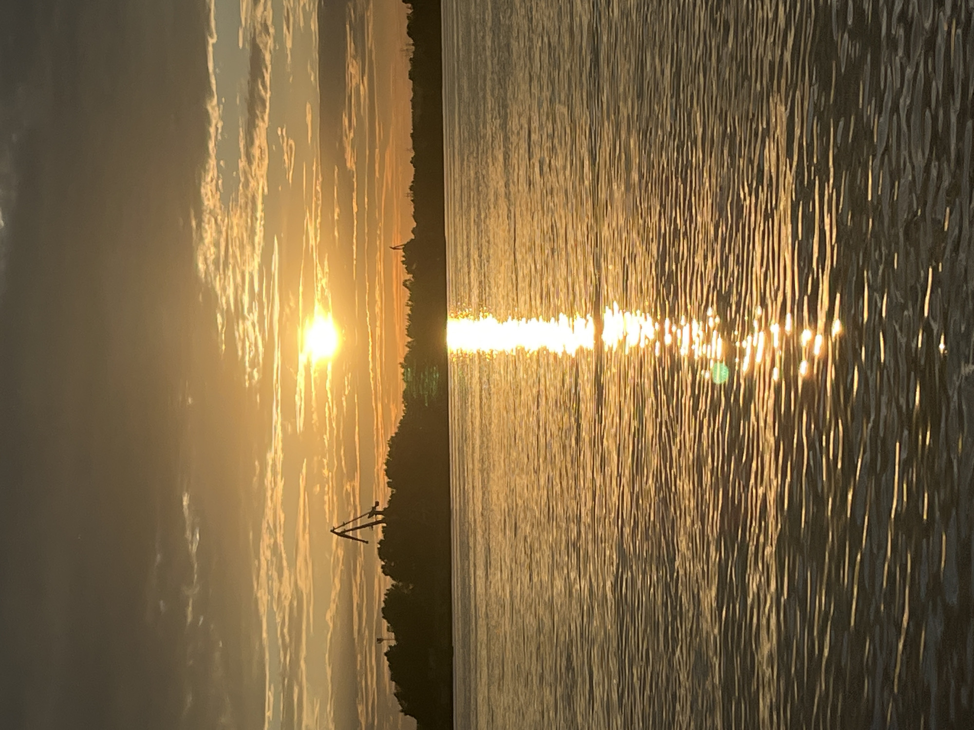 The image shows a sunset over a body of water, likely the Kama River, in Chaikovsky, Russia. The sun is low on the horizon, casting a warm, golden glow across the sky and water. The sky is filled with scattered clouds, some of which are illuminated by the setting sun. In the distance, a silhouetted treeline borders the water, and a large crane is visible against the bright sky. The water's surface is rippled, reflecting the colors of the sunset. The overall scene is serene and picturesque.