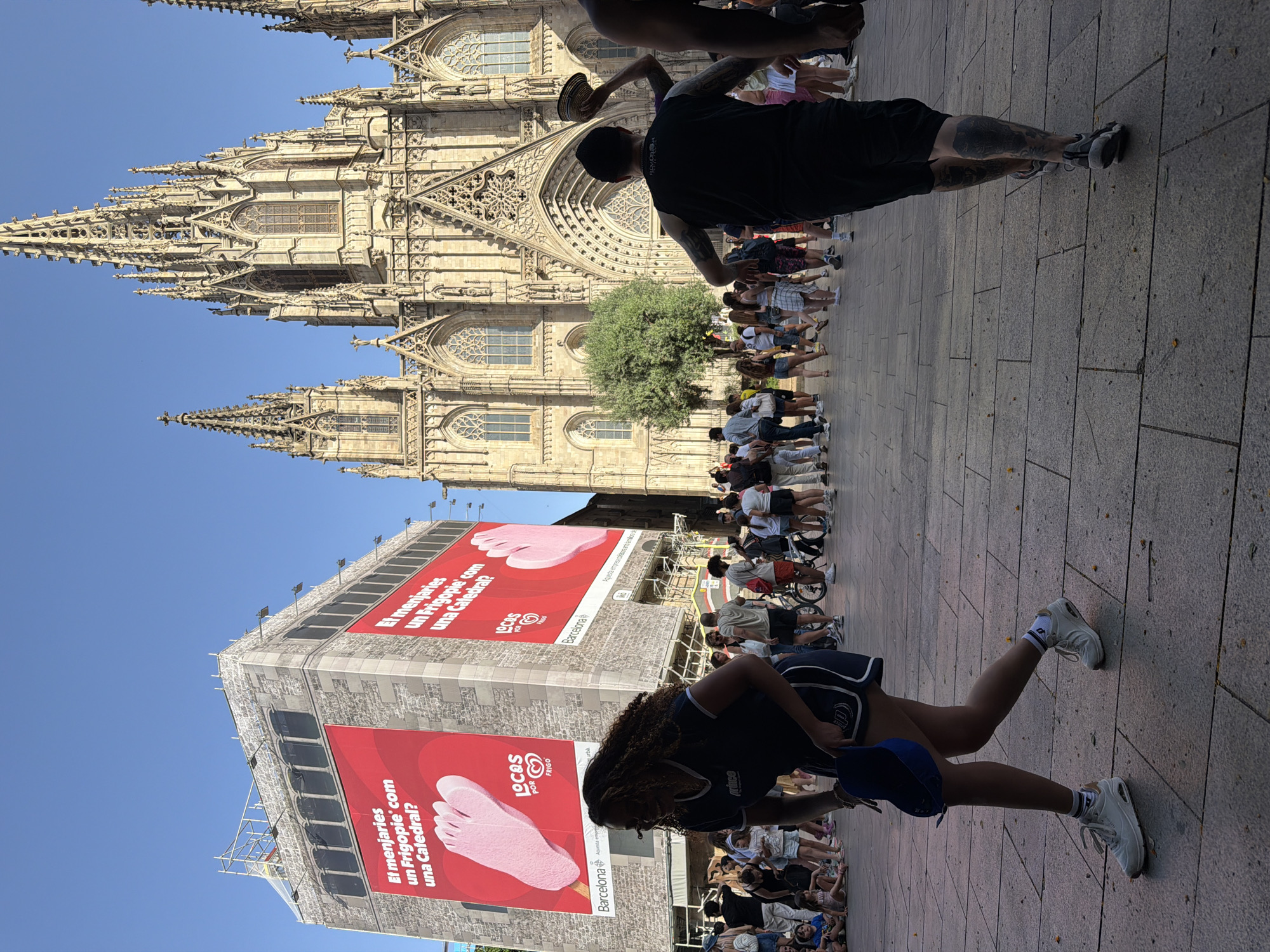 In the city of Barcelona, Spain, a bustling plaza is captured on a sunny day, with the majestic Gothic Cathedral of Barcelona dominating the background. The Cathedral's ornate spires and intricate architectural details are clearly visible against a clear blue sky. In the foreground, a large billboard plastered on a building features a bright red advertisement with images of feet and a Catalan slogan.
A woman in athletic wear with curly hair walks past the billboard, holding a blue hat. Further into the plaza, a man with tattoos on his legs walks away from the camera, holding a hat in his raised hand. Numerous other individuals are scattered throughout the plaza, some walking, others standing or sitting, enjoying the public space. The ground is paved with large, square stone tiles. In the mid-ground, a bicycle is visible, suggesting a mode of transportation for some visitors. The overall scene depicts a vibrant and active urban environment, blending historical architecture with contemporary life.
