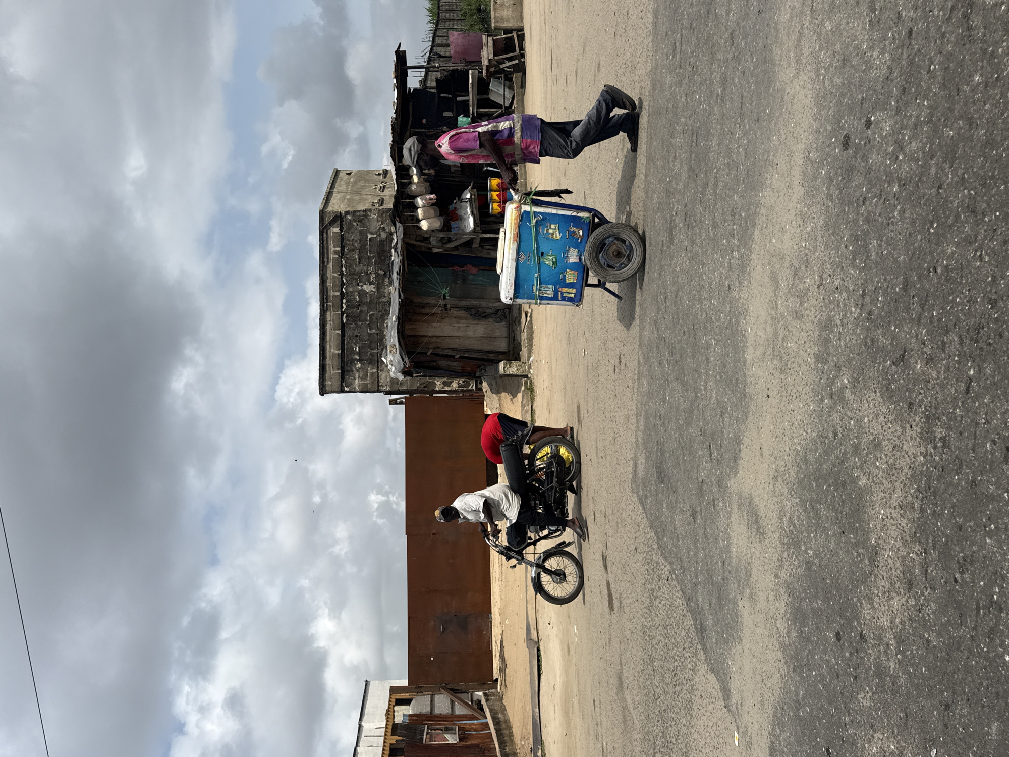A man is seen pushing a cooler on wheels down a street in Ado, Nigeria. The cooler is blue and white with colorful images of drinks on its side. He is wearing a pink and purple striped shirt and dark pants. Behind him, a man on a motorcycle, wearing a white shirt and black pants, is looking towards the camera. Another person in a red shirt is bent over the back of the motorcycle. The street appears to be unpaved in some areas. In the background, there are buildings with corrugated metal roofs and cinder block walls. The sky is cloudy.