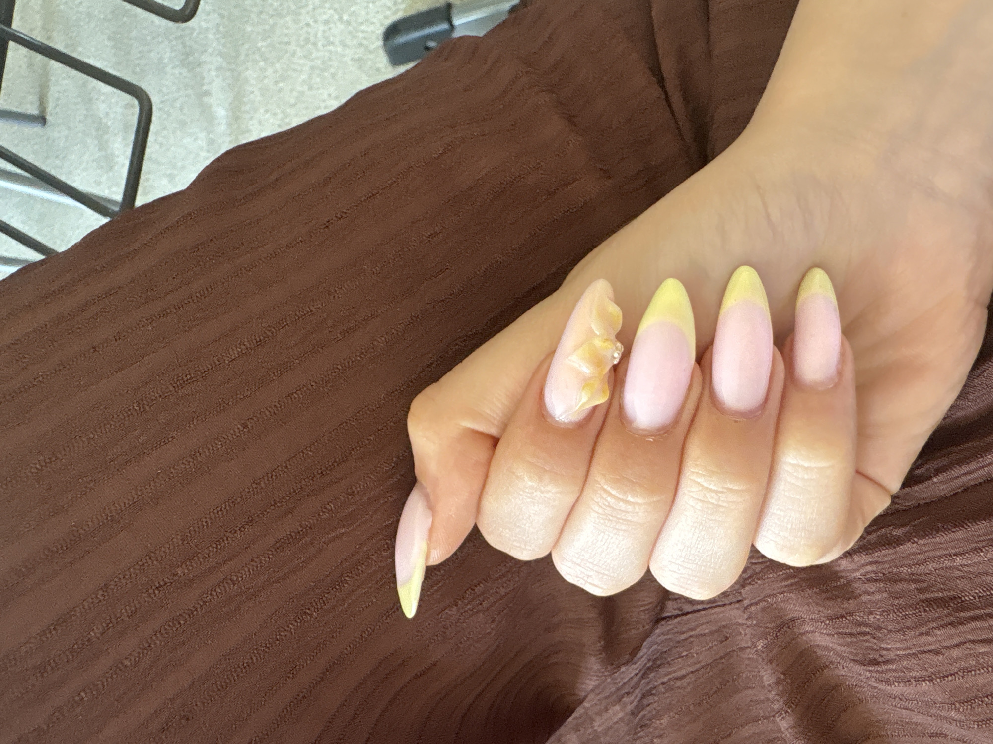 A close-up captures a human hand with long nails featuring a pastel pink base and yellow tips, arranged against a textured brown fabric. The thumb nail has an additional floral embellishment. The location is not discernible from the image.