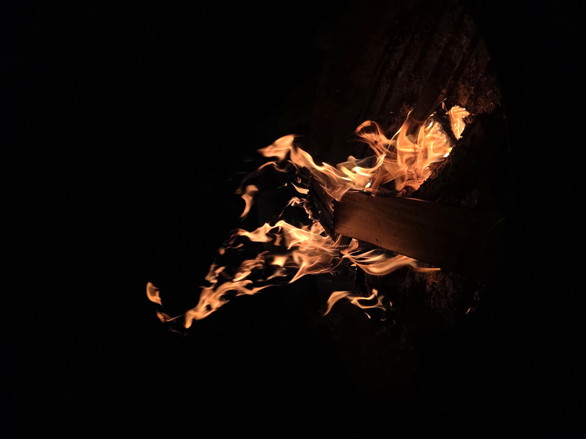 The image depicts a close-up, dimly lit shot of a campfire in the city of Строитель-нефтяник Снт, Russia. The primary subject is the fire itself, with vibrant orange and yellow flames licking upwards against a black background. The flames are dynamic and appear to be in motion, with wisps and tendrils reaching out. Within the fire, pieces of wood are visible, appearing charred and in the process of burning. The wood seems to be arranged in a way that sustains the fire. The overall impression is one of warmth and natural energy, set against the starkness of darkness. There are no people or animals visible in the scene.