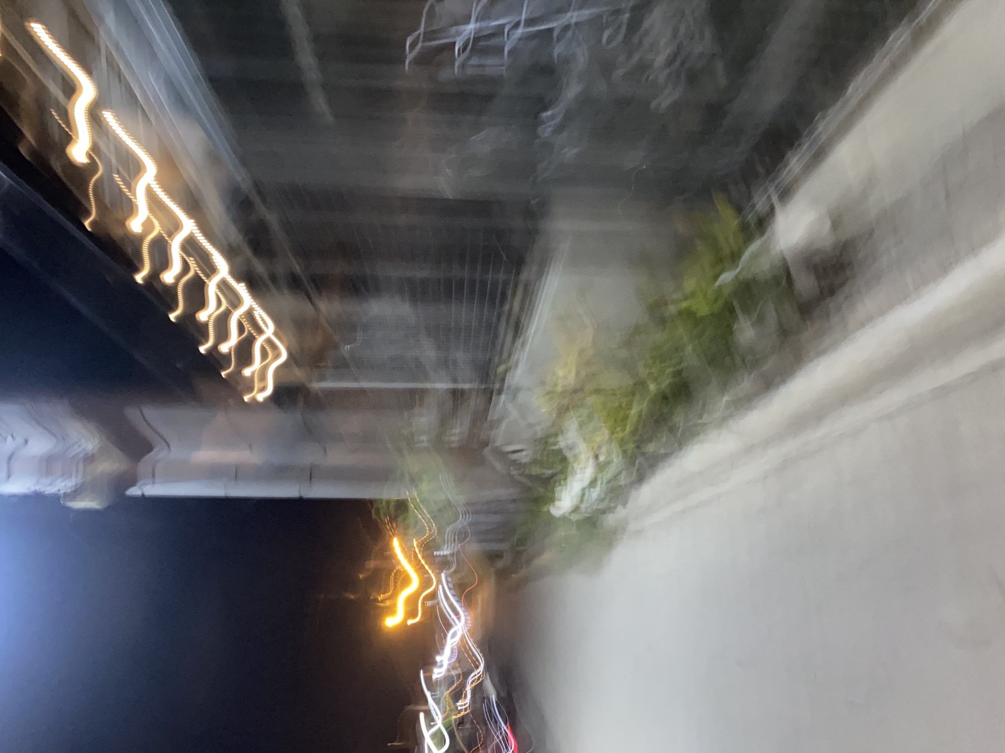 This is a forensic description of a heavily motion-blurred night photograph taken in Davao City, Philippines. The scene depicts a street or pathway, with a light-colored, paved surface occupying the lower and central foreground, receding into the distance. The primary visual elements are light trails: bright yellow-orange, wavy streaks extend horizontally across the upper right, originating from an obscured light source on an adjacent structure. On the left, multiple white and red serpentine light trails indicate the passage of vehicles, with red suggesting taillights and white suggesting headlights. A vertically oriented, blurred light-colored architectural structure, possibly a building facade or wall, is present on the right, accompanied by indistinct green vegetation at its base. Further right, a dark, vertically striped or gridded element, possibly a fence or gate, is visible. The upper left of the image features a dark, featureless night sky with a subtle luminous haze at the very top. No discernible individuals or specific objects are clearly identifiable, with the entire composition conveying a sense of rapid movement through a low-light urban environment.