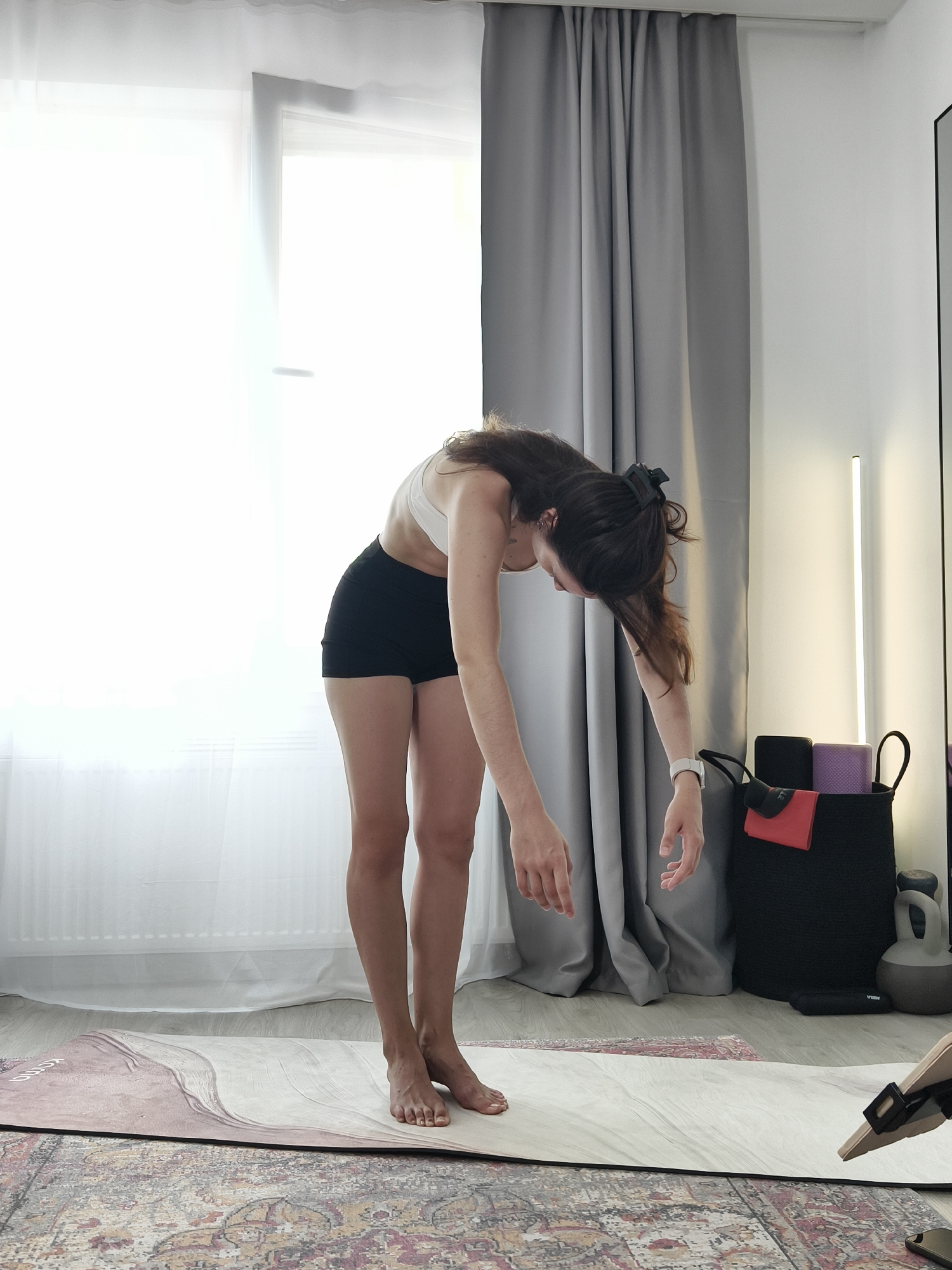 A woman is performing a yoga pose indoors. She is standing on a yoga mat in a room with light-colored wooden floors and white walls. She is wearing a white sports bra and black shorts. Her body is bent forward at the waist, with her arms hanging loosely towards the floor. Her hair is long and dark brown, pulled back with a clip. In the background, there are grey curtains, a large window with white sheer curtains, and a black basket containing yoga blocks and a red towel. A kettlebell and other fitness equipment are visible on the floor. The scene is well-lit, with natural light streaming in from the window. The image does not provide any information about the location in Tyumen, Russia.
