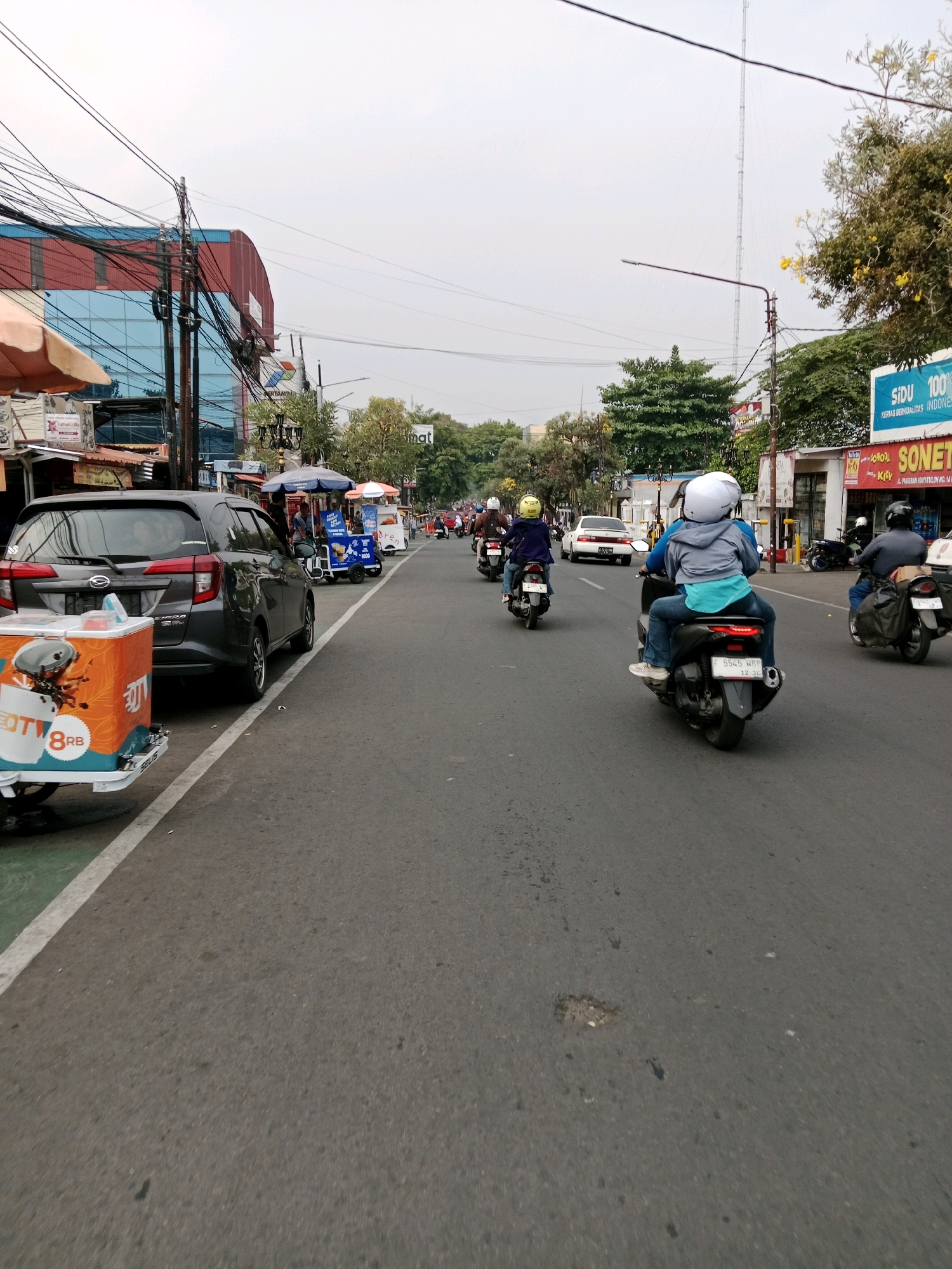 The image depicts a street scene in Cianjur, Indonesia, likely on a cloudy day. The perspective is from a low angle, possibly from a moving vehicle. A dark gray car is parked on the left side of the road, with a cart displaying an orange and white advertisement for "OTY 8RB" attached to its rear. Further down the road, several motorcycles are visible, some with riders wearing helmets and jackets. On the right side of the street, there are shops with awnings and signage, including a blue sign that reads "SIDU 100%" and "SONET". Trees line the street, and utility wires are strung overhead, creating a cluttered visual in the sky. The road surface appears to be asphalt with some minor imperfections.