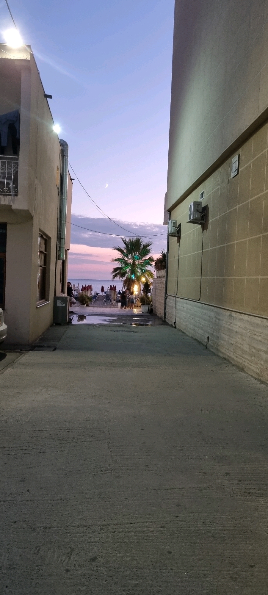 The image depicts a narrow pathway between two buildings, leading towards a beach scene at dusk. The pathway is paved with concrete and shows signs of moisture. On the left, a light-colored building has a balcony with laundry hanging and an external pipe. On the right, a larger, tan-tiled building features two air conditioning units.
The pathway opens up to a beach area with rows of umbrellas and lounge chairs. A palm tree, illuminated with green lights, stands prominently in the center of the beach area. Several people are visible near the palm tree and on the beach, some appearing to be walking or standing. The sky is a gradient of light blue and pink, with a crescent moon visible. The sea is in the background, and it appears calm. The scene suggests a coastal town or resort area.