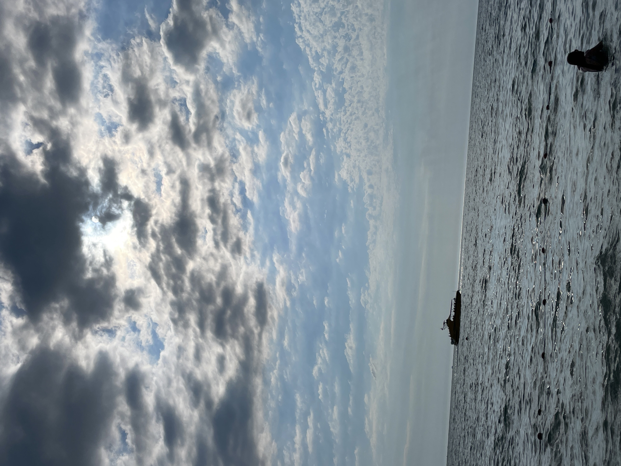 The image depicts a seascape with a boat on the horizon and a person in the foreground. The sky is filled with dramatic clouds, with the sun partially obscured behind them. The water is choppy, reflecting the light and the sky. In the distance, a yellow boat with people on board is sailing across the sea. In the foreground, a person is partially submerged in the water, facing away from the camera. A line of buoys is visible in the water between the person and the boat. The scene suggests a day at the beach or a boat tour in Sochi, Russia.