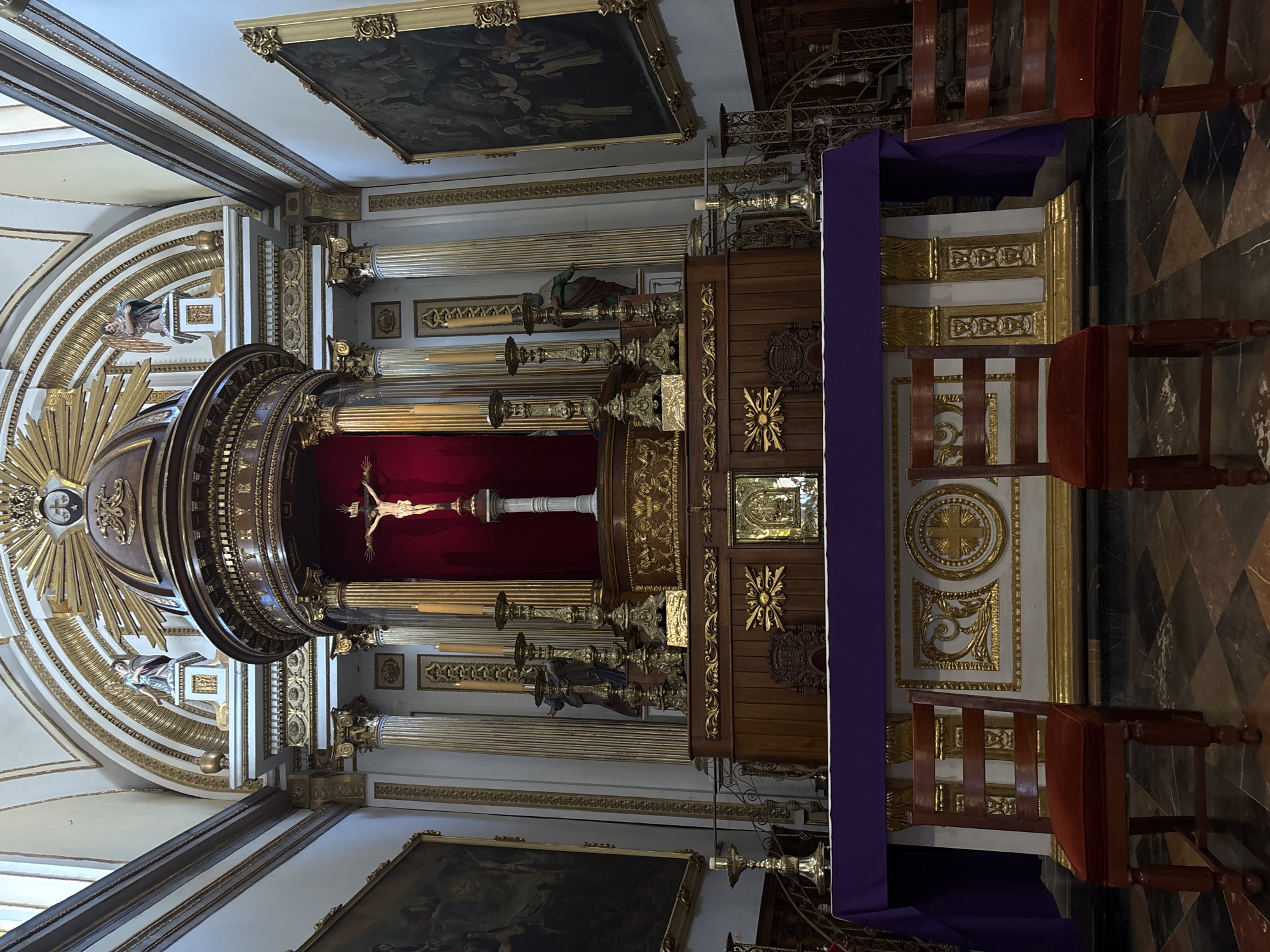 The image depicts the interior of a church altar in Puebla, Mexico. The altar features a crucifix against a red backdrop, flanked by ornate gold and wooden decorations. Statues are visible on either side of the crucifix. A purple cloth drapes over the front of the altar. Two wooden chairs are positioned in front of the altar. The church's architecture includes gold detailing, paintings on the ceiling, and a patterned floor.
