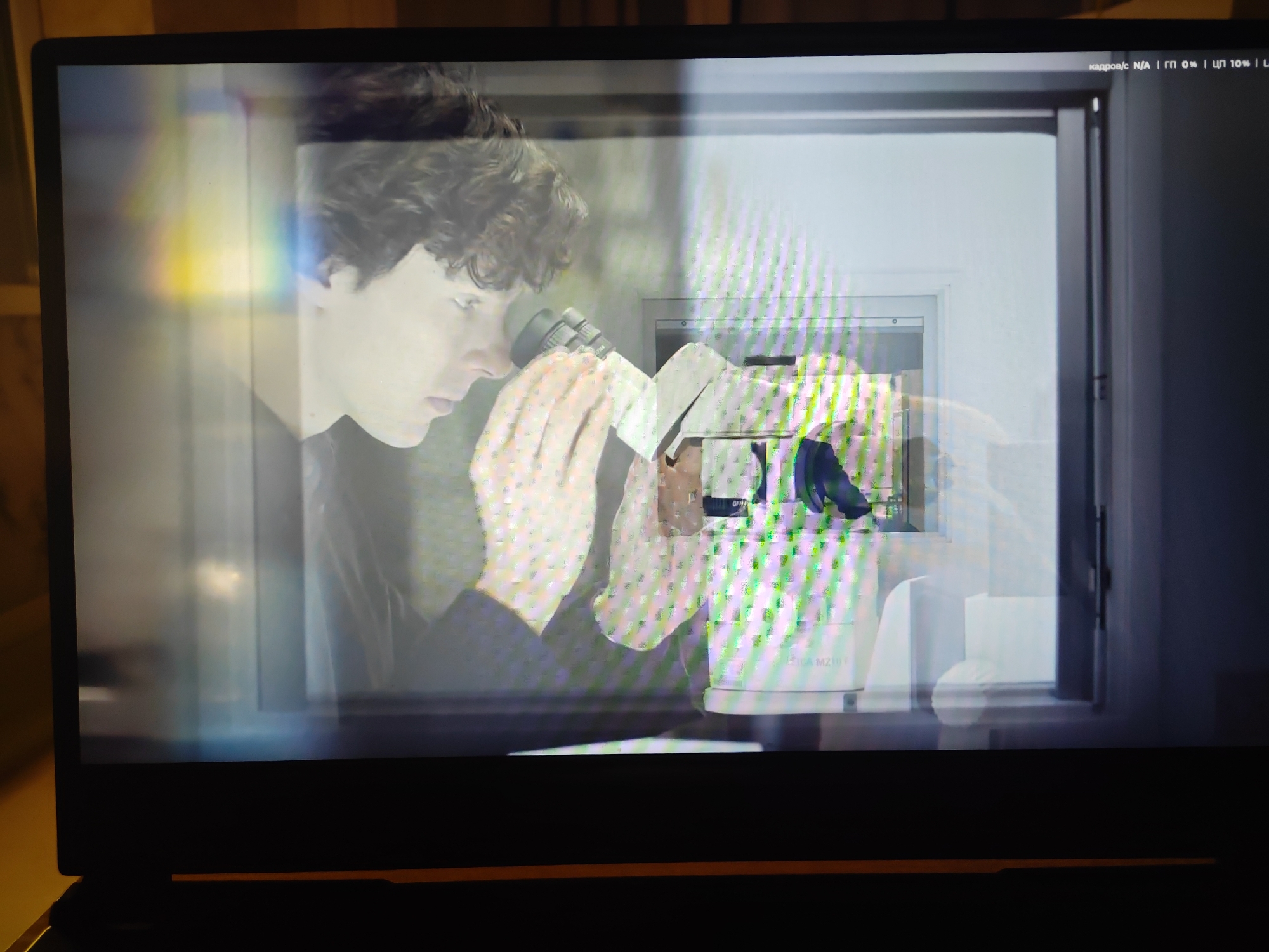 The image shows a close-up of a laptop screen displaying a scene with a young person looking through a microscope. The person has curly brown hair and is wearing a dark shirt. They are holding a white cloth to the microscope. The background appears to be a laboratory or research setting, with white equipment and shelves visible. There are digital overlays on the screen, including Russian text at the top right corner which translates to "frames/s: N/A | FPS 0% | DPI 10% | L". The screen itself has some color distortion and pixelation, particularly on the right side, suggesting a technical issue with the display or the video being played. There are also light reflections on the screen, with a rainbow-like effect on the left side. The laptop is black and appears to be sitting on a desk or table, with a hint of yellow furniture in the background. The scene does not provide any specific information or visual cues to indicate a location in Krasnodar, Russia, or any other specific city.