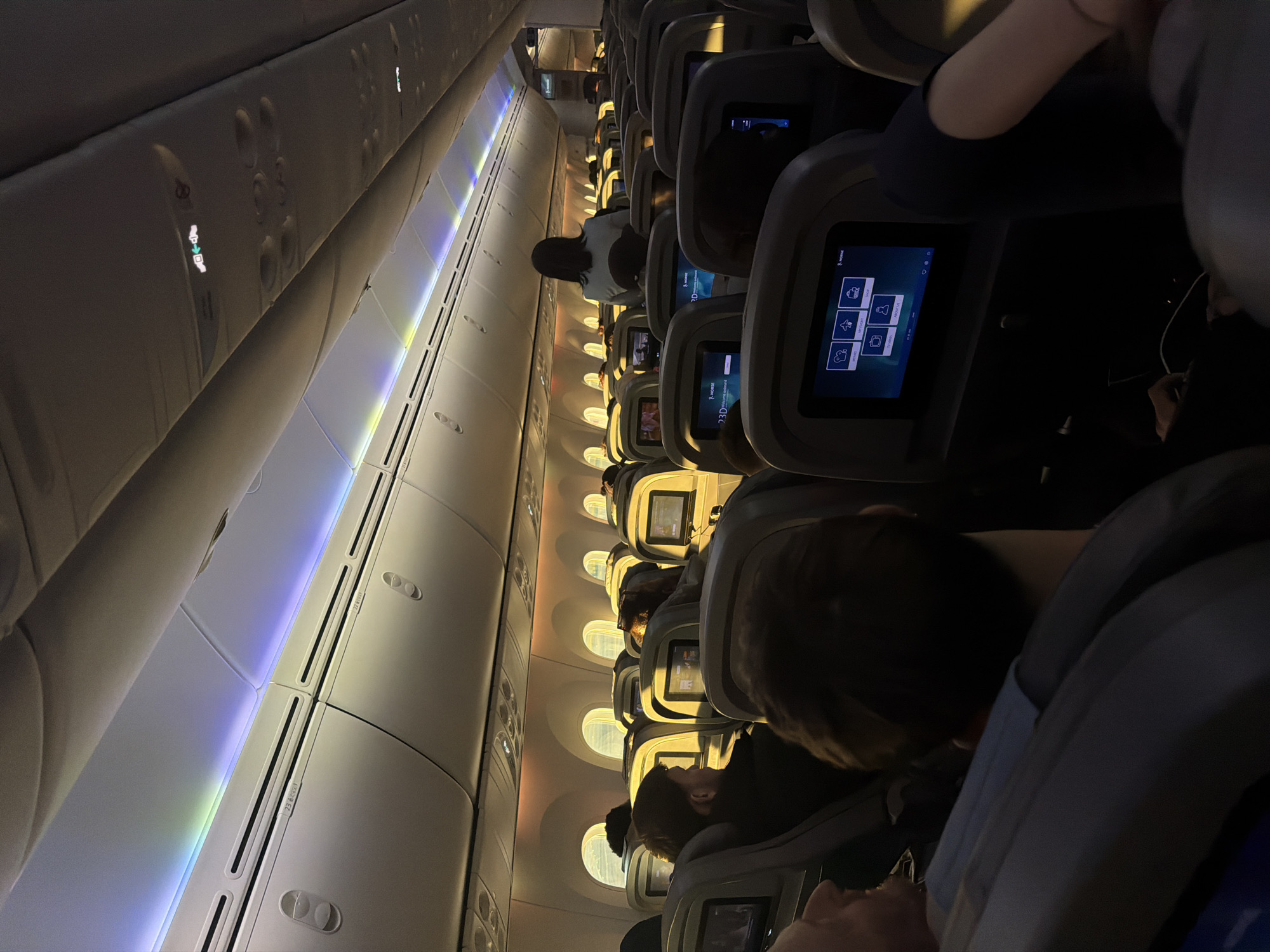 The image shows the interior of an airplane cabin. Rows of seats are visible, most of which are occupied by passengers. Individual screens are attached to the back of each seat, displaying various images and interfaces. The overhead compartment area is visible, along with illuminated panels with air vents. The general lighting is dim, with accents from the overhead cabin lights. The scene depicts passengers in an airplane cabin during a flight.