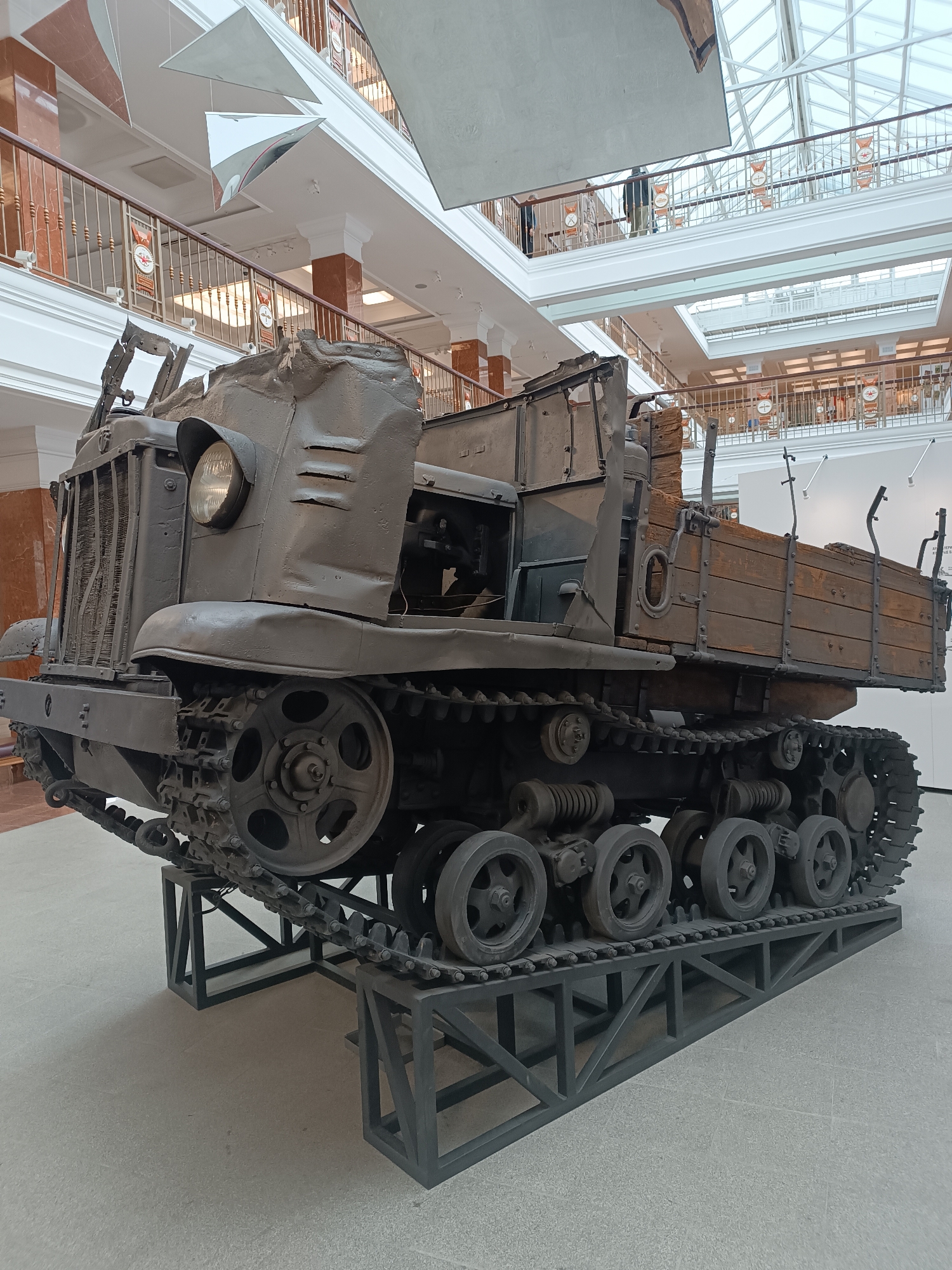 A drab, olive-green, tracked vehicle, likely a museum exhibit, is positioned on a metal display stand in the center of a modern, multi-level building with a glass ceiling. The vehicle, which appears to be a vintage military truck or tractor, is a dominant subject in the foreground. It has a large, round headlight on the front, a metal grille, and a wooden cargo bed at the rear. The vehicle is elevated on a dark grey metal frame constructed of triangular and rectangular supports. The environment suggests an indoor exhibition hall, with balconies and railings visible in the background. The floor is a light-colored, speckled material. The lighting is bright and diffused, indicating overhead illumination. There are no people directly interacting with the vehicle, though individuals can be seen on the upper levels of the building, observing the exhibits. The overall scene is static and showcases the historical artifact. This vehicle is located in the Museum of Military Equipment UMMC in Verkhnyaya Pyshma, Russia.