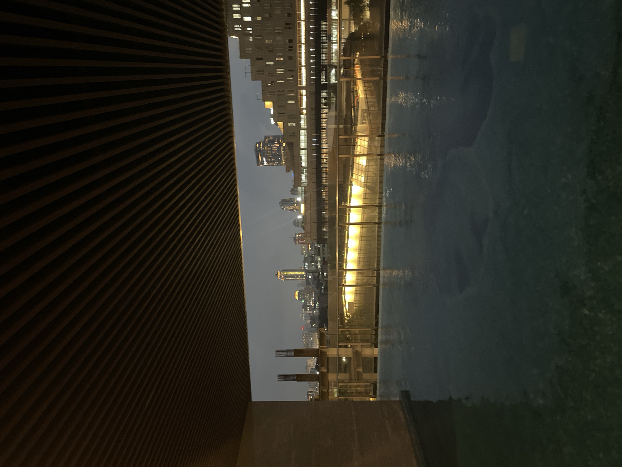 The image captures a twilight view of a city skyline, likely Central Jakarta, Indonesia, from an elevated vantage point, possibly a rooftop or balcony. In the foreground, a calm body of water, likely a swimming pool or reflecting pool, occupies the lower half of the frame. The water's surface reflects the lights of the surrounding buildings, creating a shimmering effect.
Above the water, a sleek, modern architectural element, possibly a ceiling or overhang constructed of dark wooden slats, frames the view of the city. To the left, a substantial concrete structure, part of the building from which the photo is taken, partially obstructs the view.
The cityscape in the background is a dense collection of buildings, illuminated by artificial lights. Various skyscrapers and mid-rise structures of different architectural styles are visible, indicating a bustling urban environment. Some buildings are lit with warm, yellow lights, while others display cooler, white illumination. The sky is overcast and dark, typical of a city at dusk or during a cloudy evening. The overall impression is one of urban sophistication and serene evening ambiance.