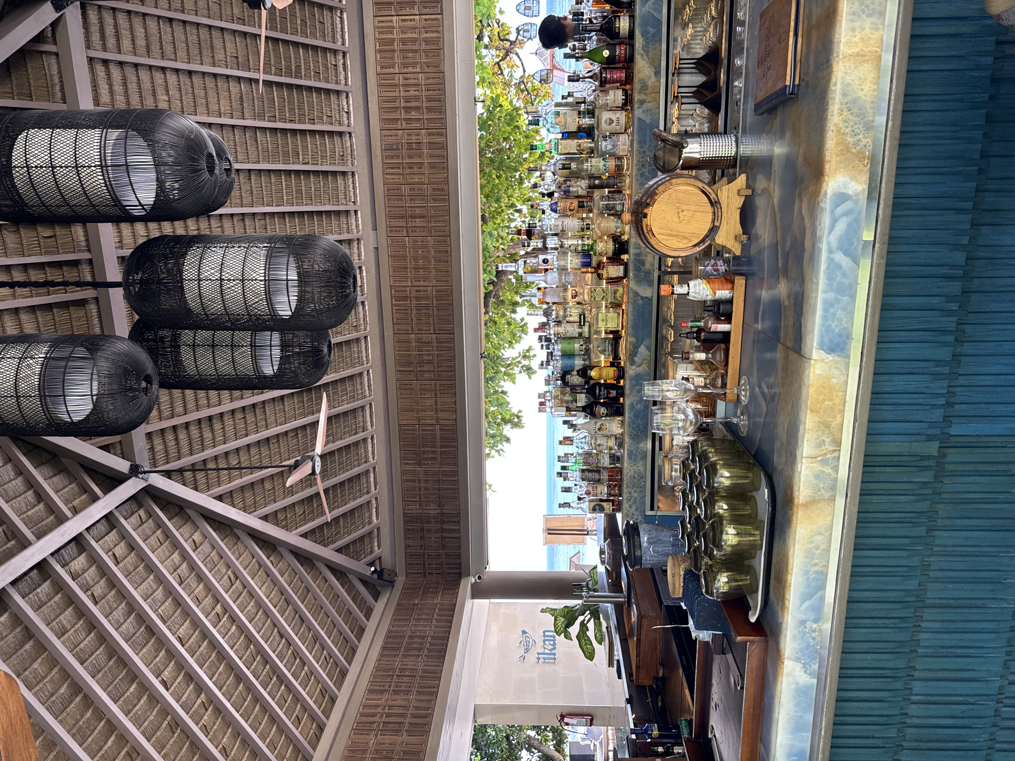 The image shows an indoor/outdoor bar area in Nusa Dua, Indonesia. The ceiling has a thatched roof supported by wooden beams with three decorative, black, wicker-like light fixtures hanging from it. A ceiling fan is also visible. The bar itself is well-stocked with numerous bottles of liquor displayed on shelves. A small wooden barrel is on the counter. A collection of glasses on a tray sits nearby. A view of the ocean can be seen through an opening behind the bar. The wall adjacent to the bar features the word "ikan" with a small graphic.