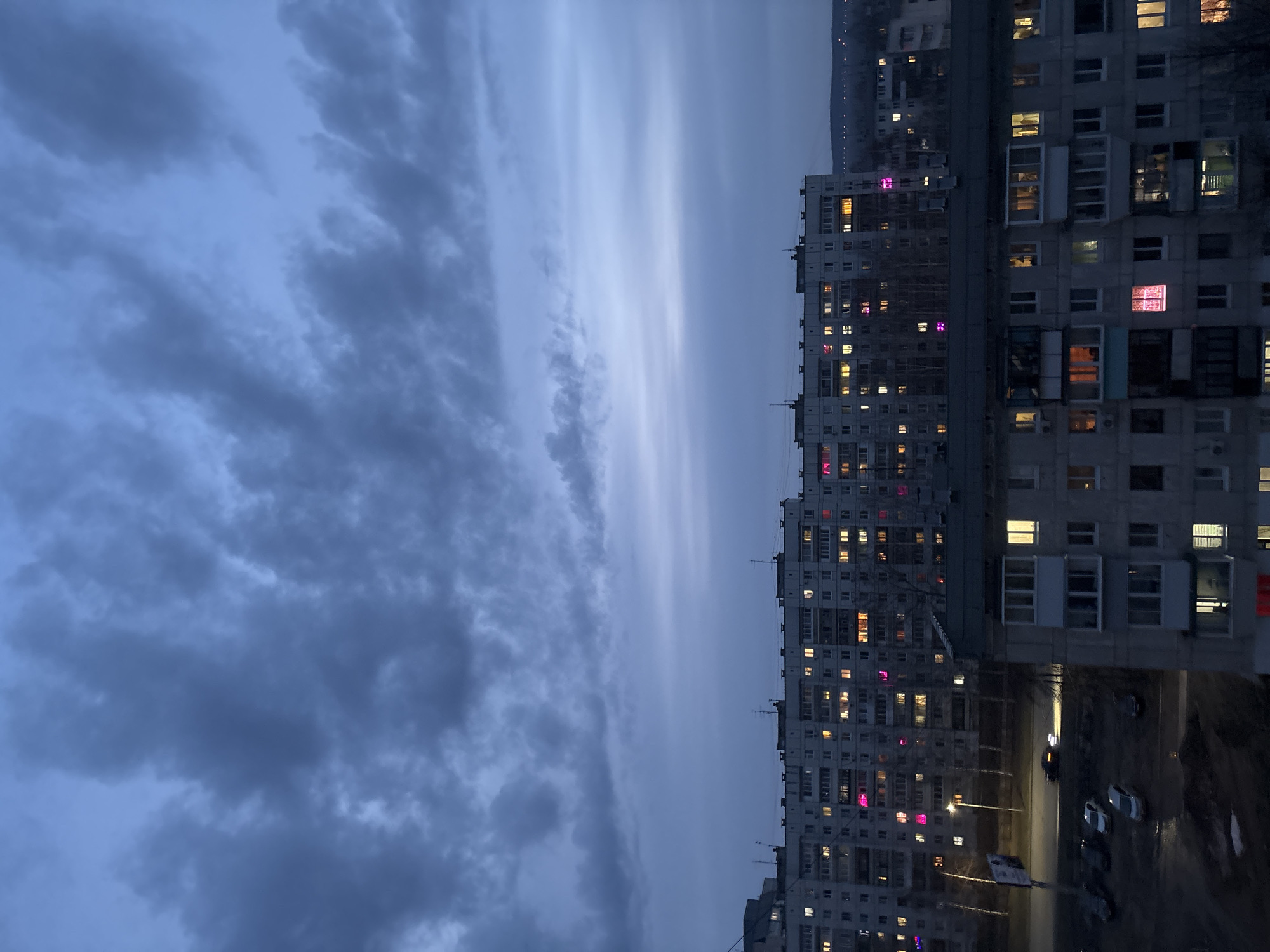 The image presents an evening scene in what appears to be a residential area, possibly in or around the city of Chaikovsky, Russia. The sky is heavily overcast with dark, textured clouds dominating the left side of the frame. A row of tall apartment buildings stands on the right, their facades punctuated by numerous windows, many of which are illuminated, indicating nighttime activity. Some windows display distinct pink lighting. Streetlights cast a warm glow on a street in the foreground, where several parked cars are visible. The overall atmosphere is subdued and urban.